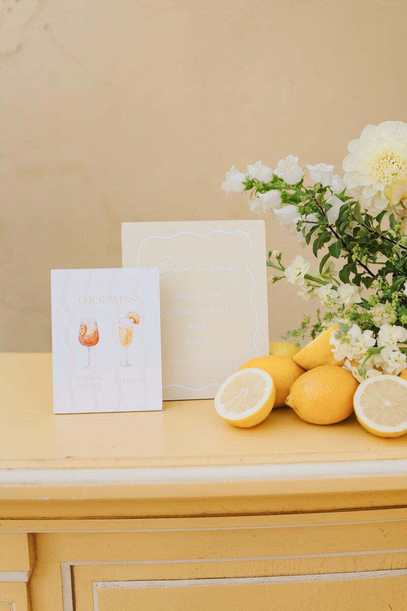 Cocktail bar menu with Aperol Spritz illustration, fresh lemons, and white floral arrangement on yellow dresser