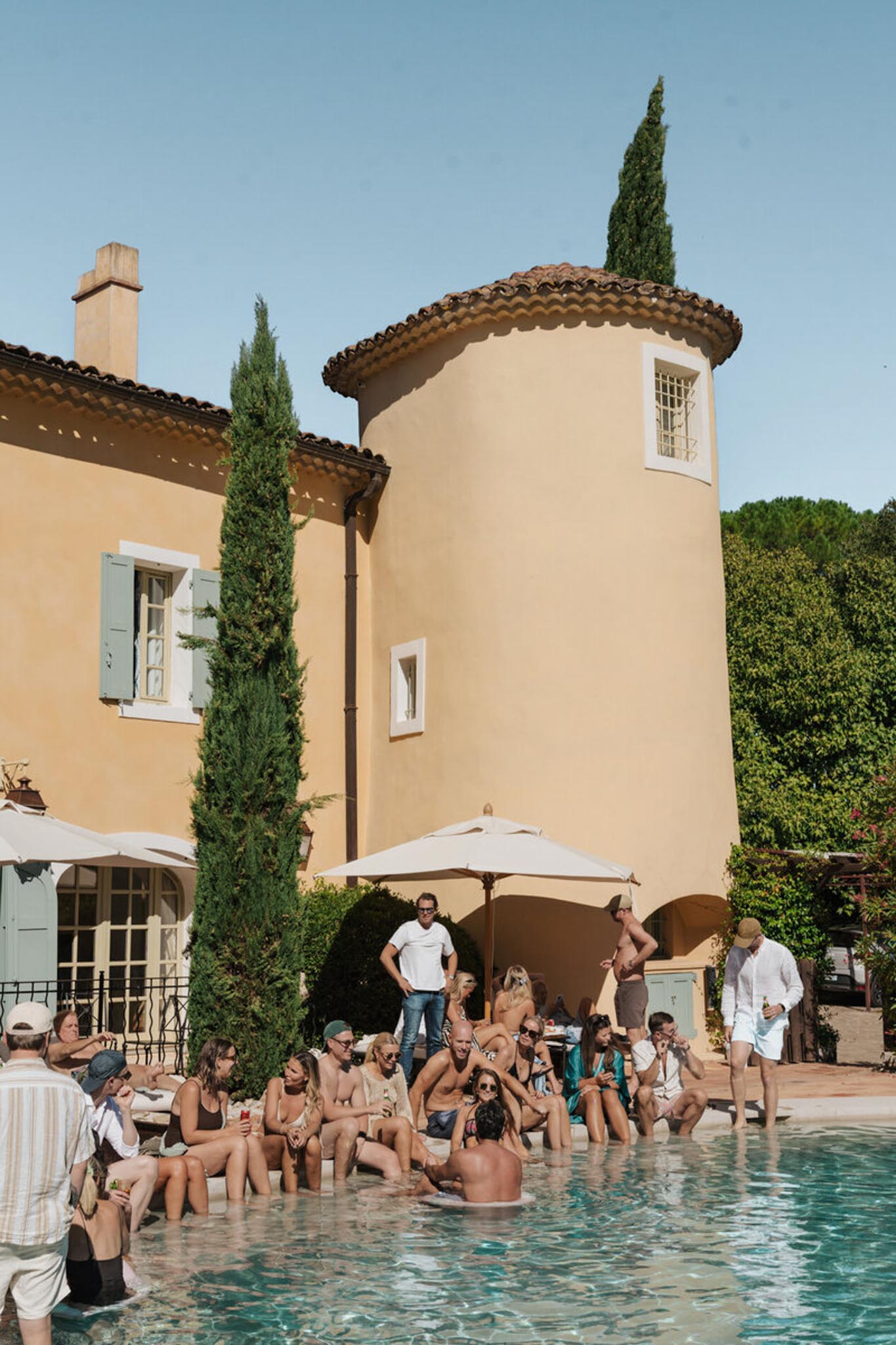 Poolside gathering twenty guests in swimwear at Provencal villa with ochre walls round tower and cream umbrellas