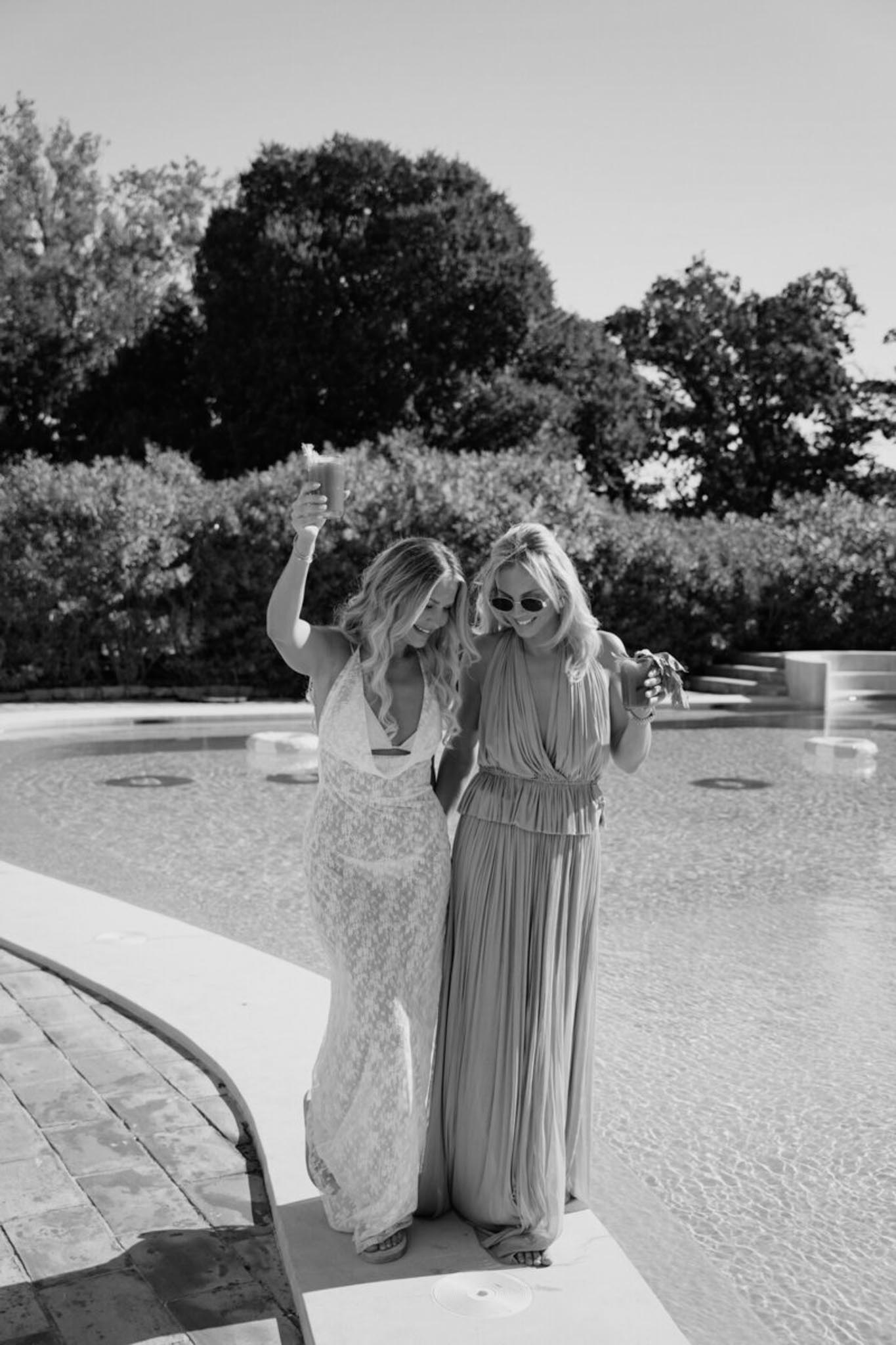 Black and white portrait of two women in maxi dresses raising drinks at poolside during cocktail hour