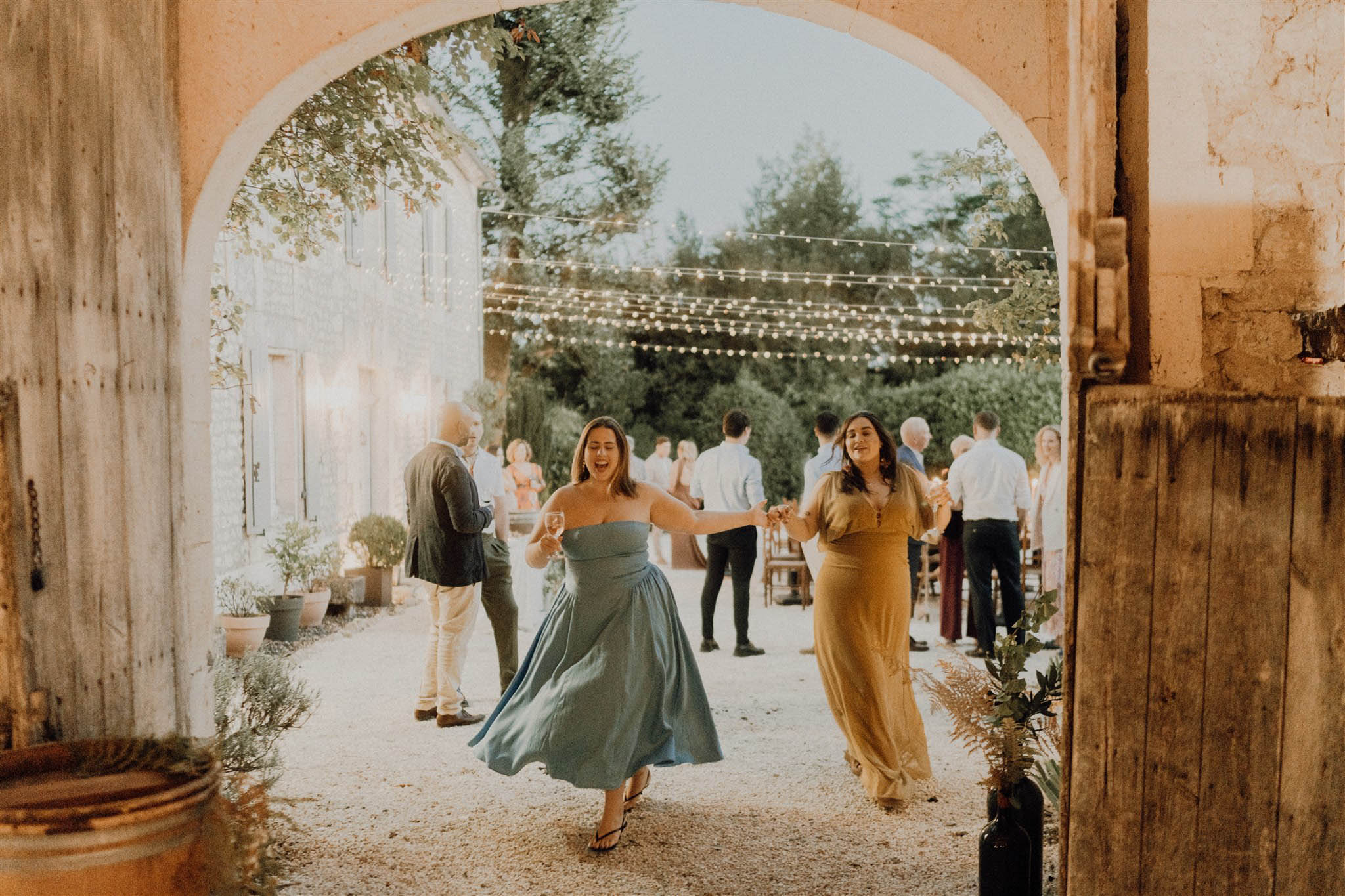 Two guests dancing through stone archway at dusk reception with string lights in French courtyard