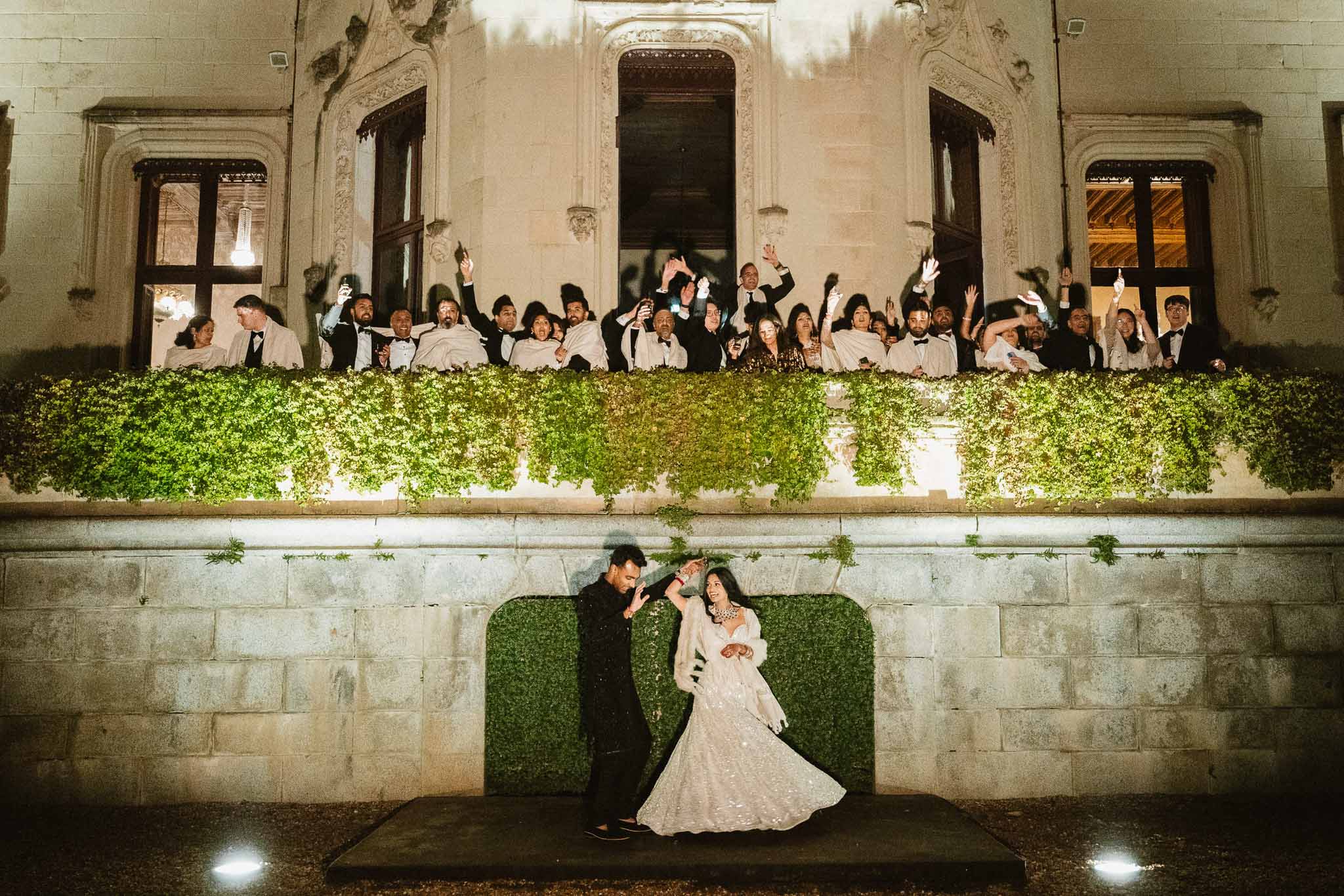 Bride and groom on stage with guests celebrating from historic stone building balcony at evening reception