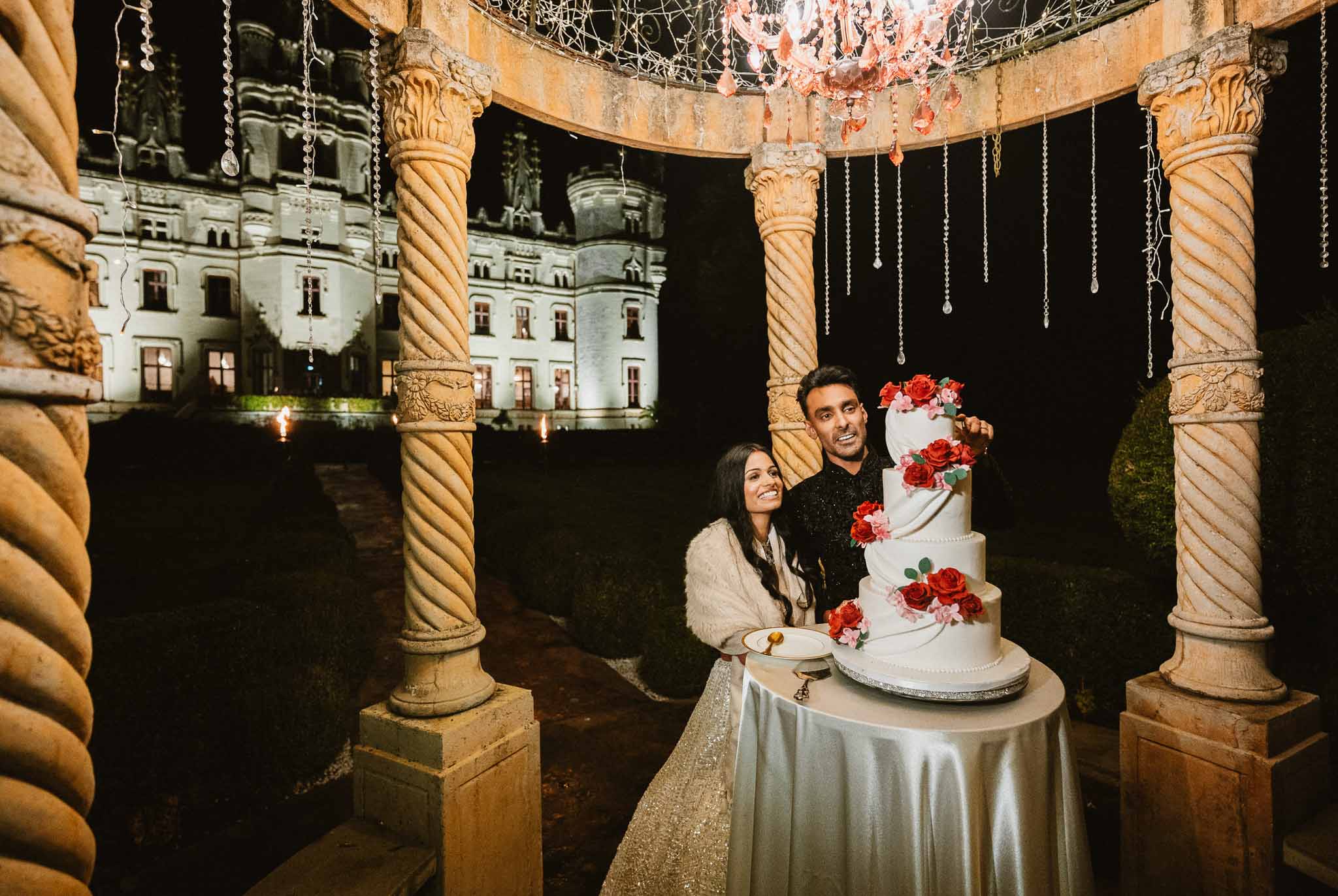 Bride and groom cut four-tier rose cake under crystal chandelier rotunda with illuminated chateau behind