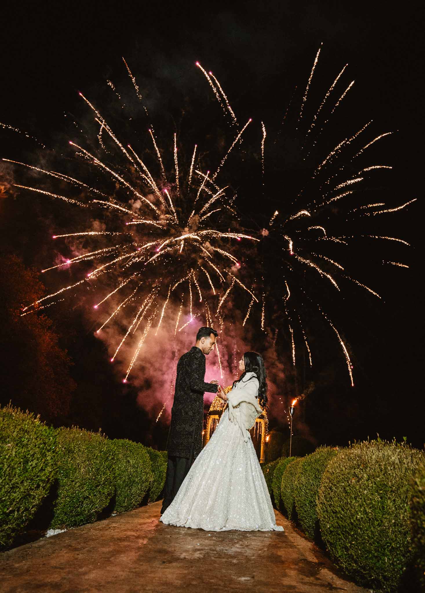 Bride and groom watching fireworks display in formal garden at night reception