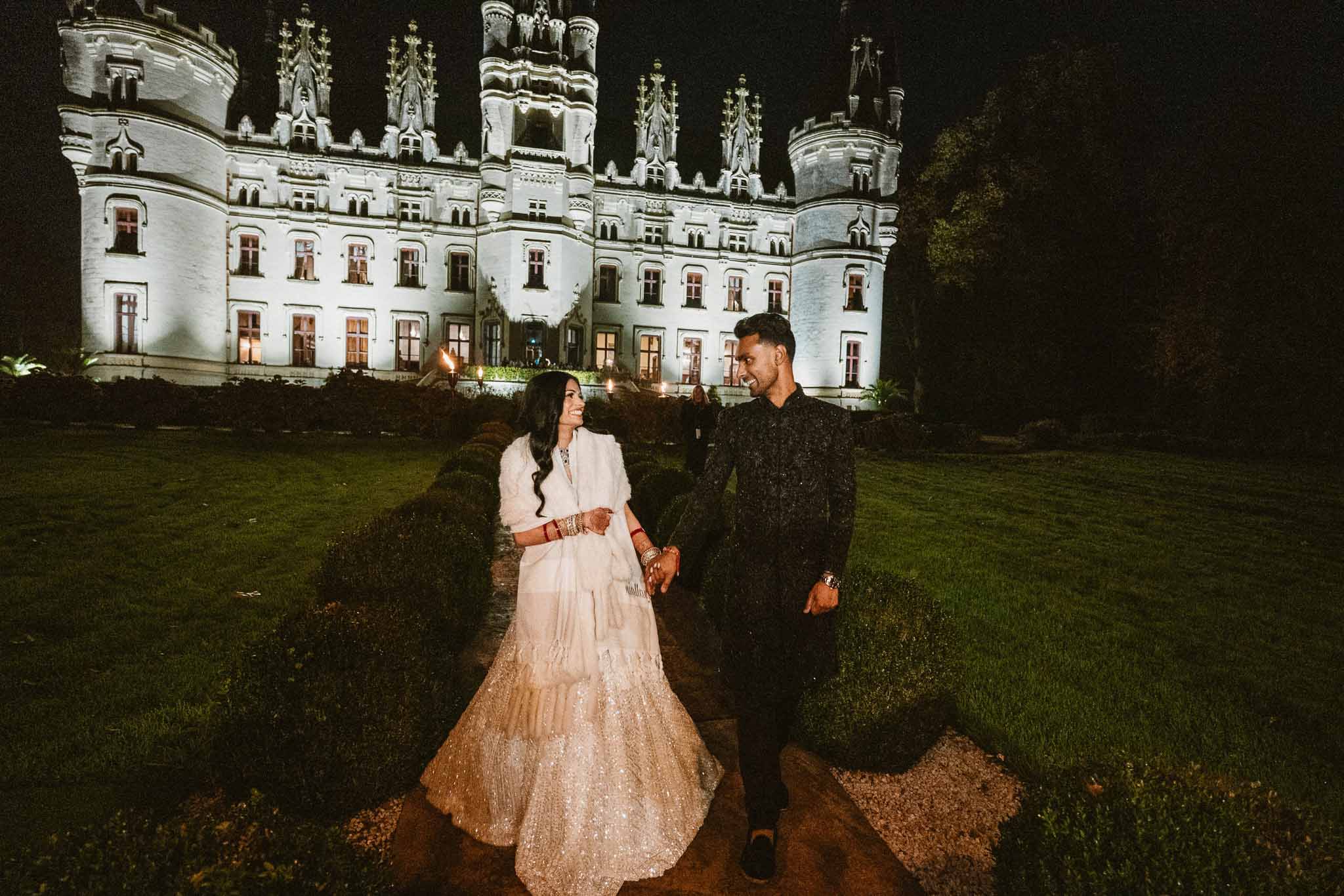 Bride and groom walking hand-in-hand in front of illuminated Gothic Revival castle during evening portraits