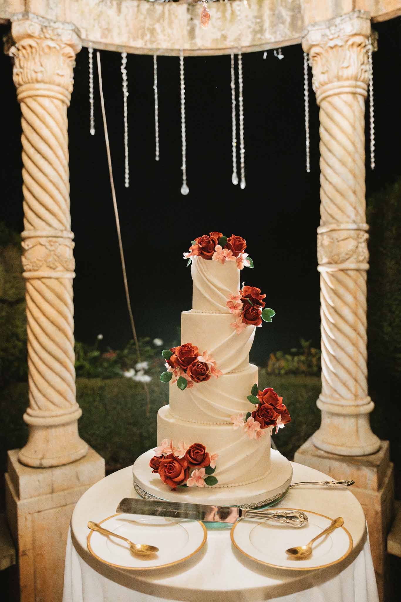 Five-tiered ivory wedding cake with burgundy roses during cake cutting at outdoor evening reception