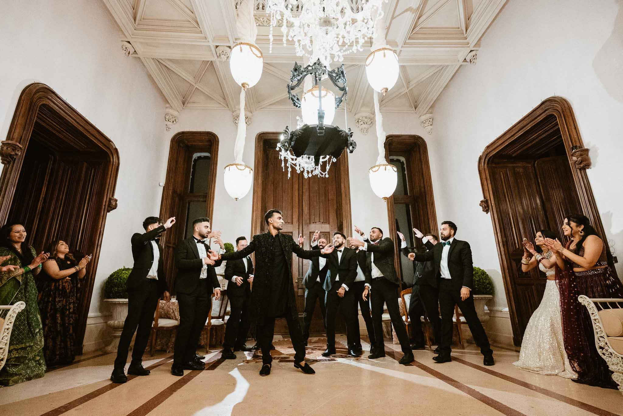 Groom and wedding party celebrating in ornate ballroom with crystal chandelier and classical architecture