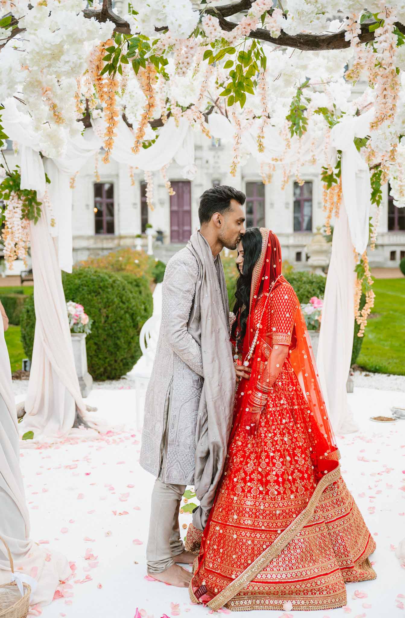 Bride and groom in traditional South Asian attire under floral archway in formal courtyard