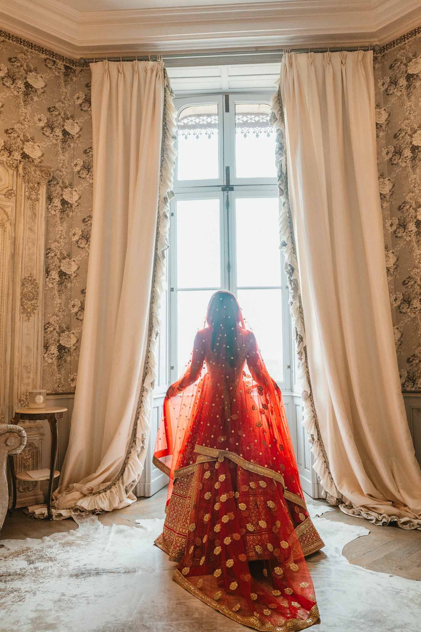 Bride in red lehenga facing window in classical interior room during getting ready