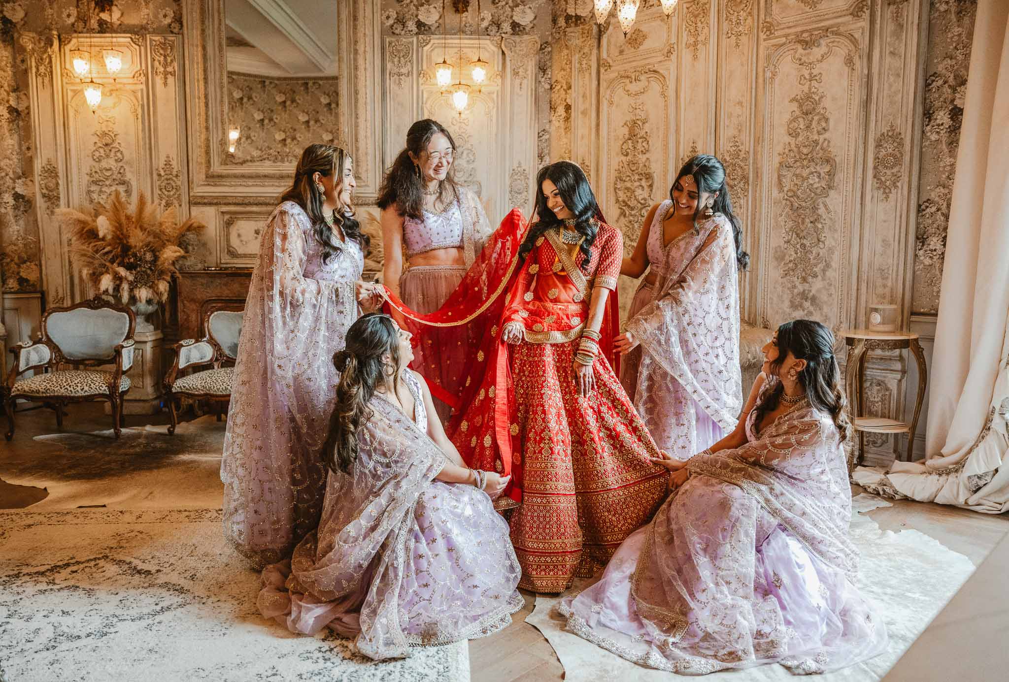 Bride in red lehenga with bridesmaids during getting ready moment in ornate ballroom interior
