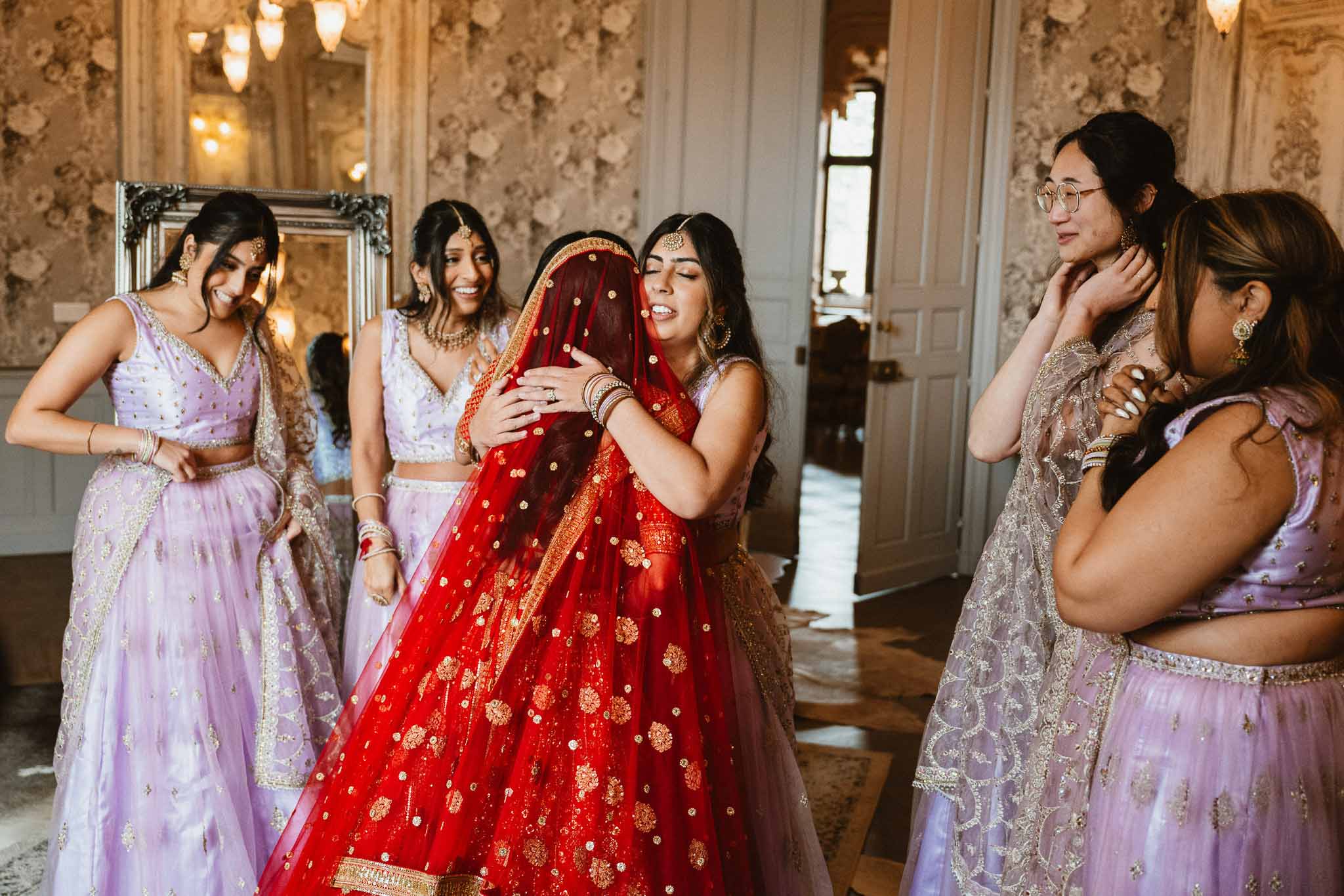 Bride in red lehenga with bridesmaids in lavender getting ready in elegant interior room