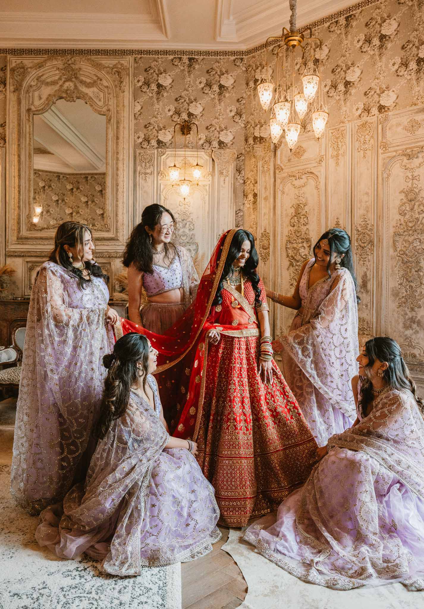 Bride in red lehenga with bridesmaids during getting ready moment in ornate ballroom
