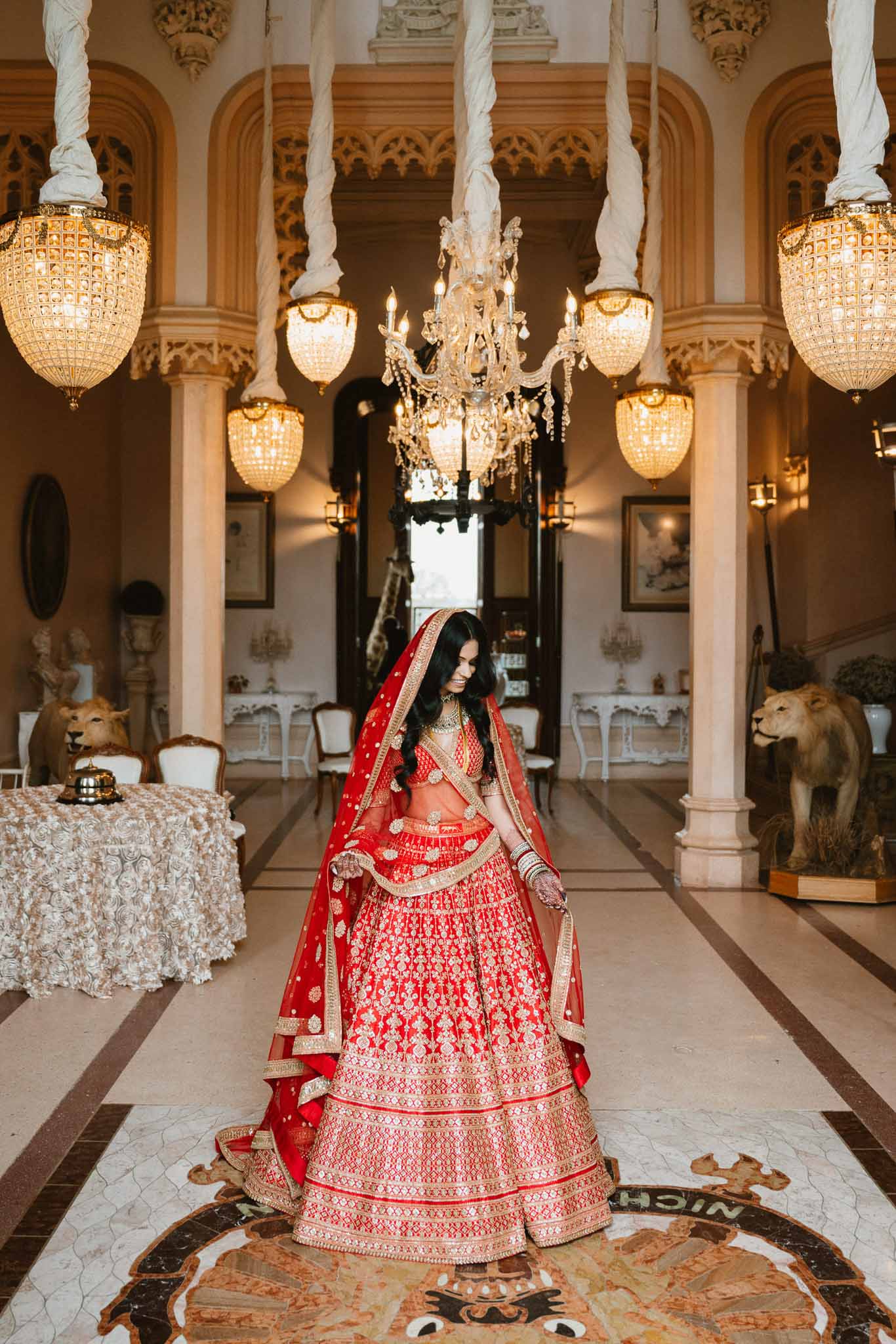 Indian bride in red lehenga standing in ornate ballroom with crystal chandeliers and neoclassical architecture