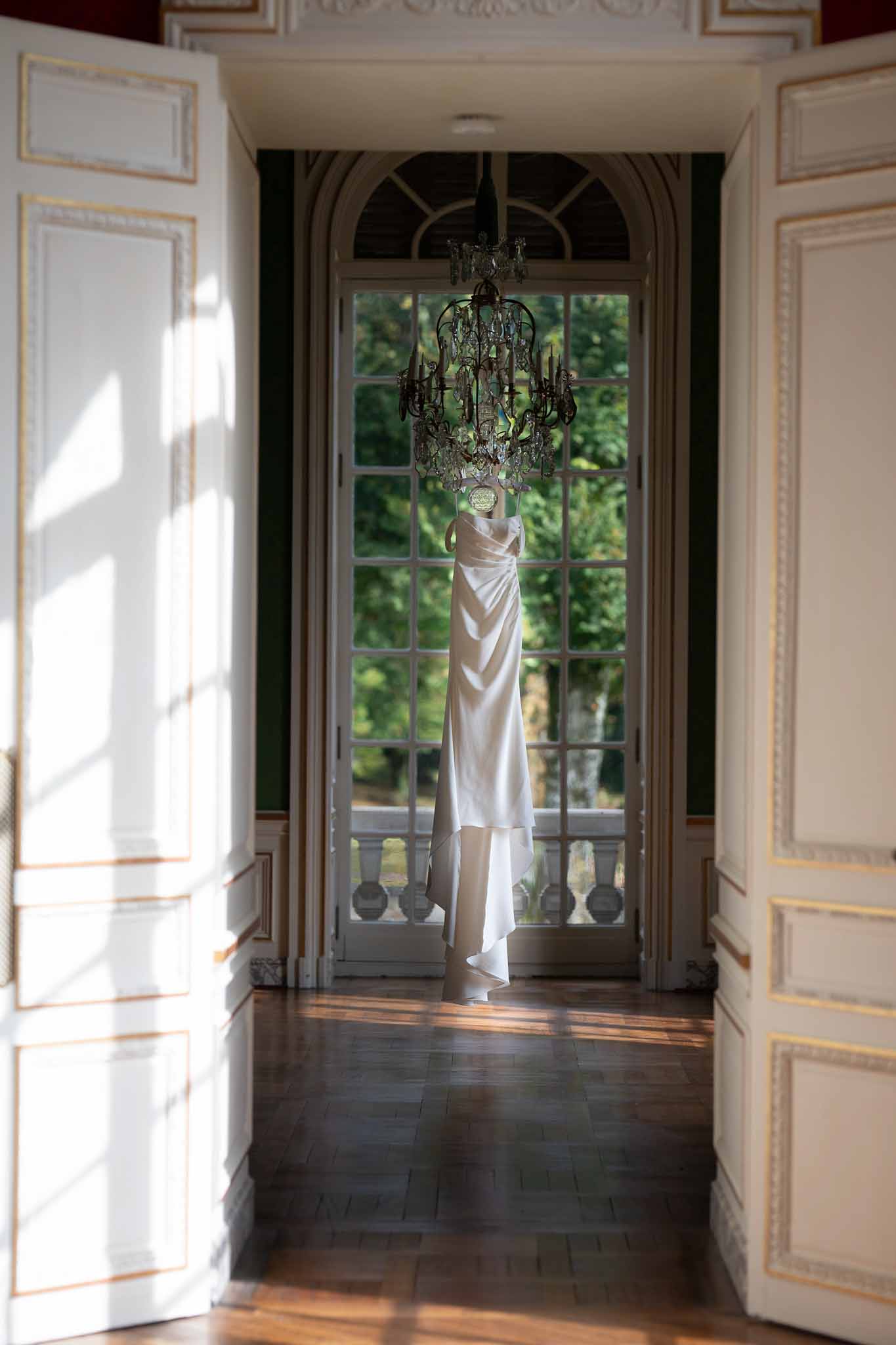 Wedding dress hanging from crystal chandelier in classical venue hallway with arched windows