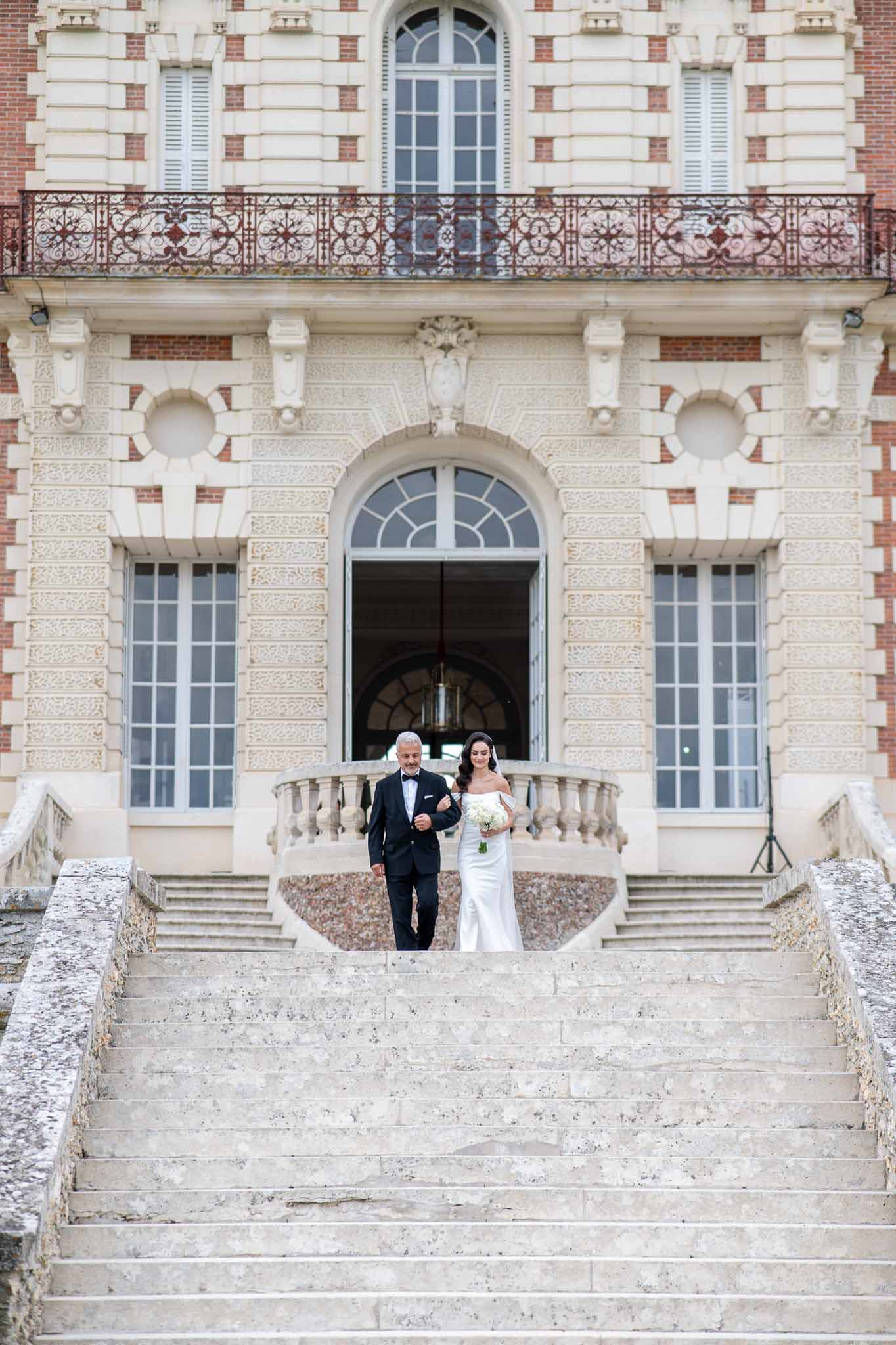 Bride and father walking down stone staircase at classical château during wedding procession