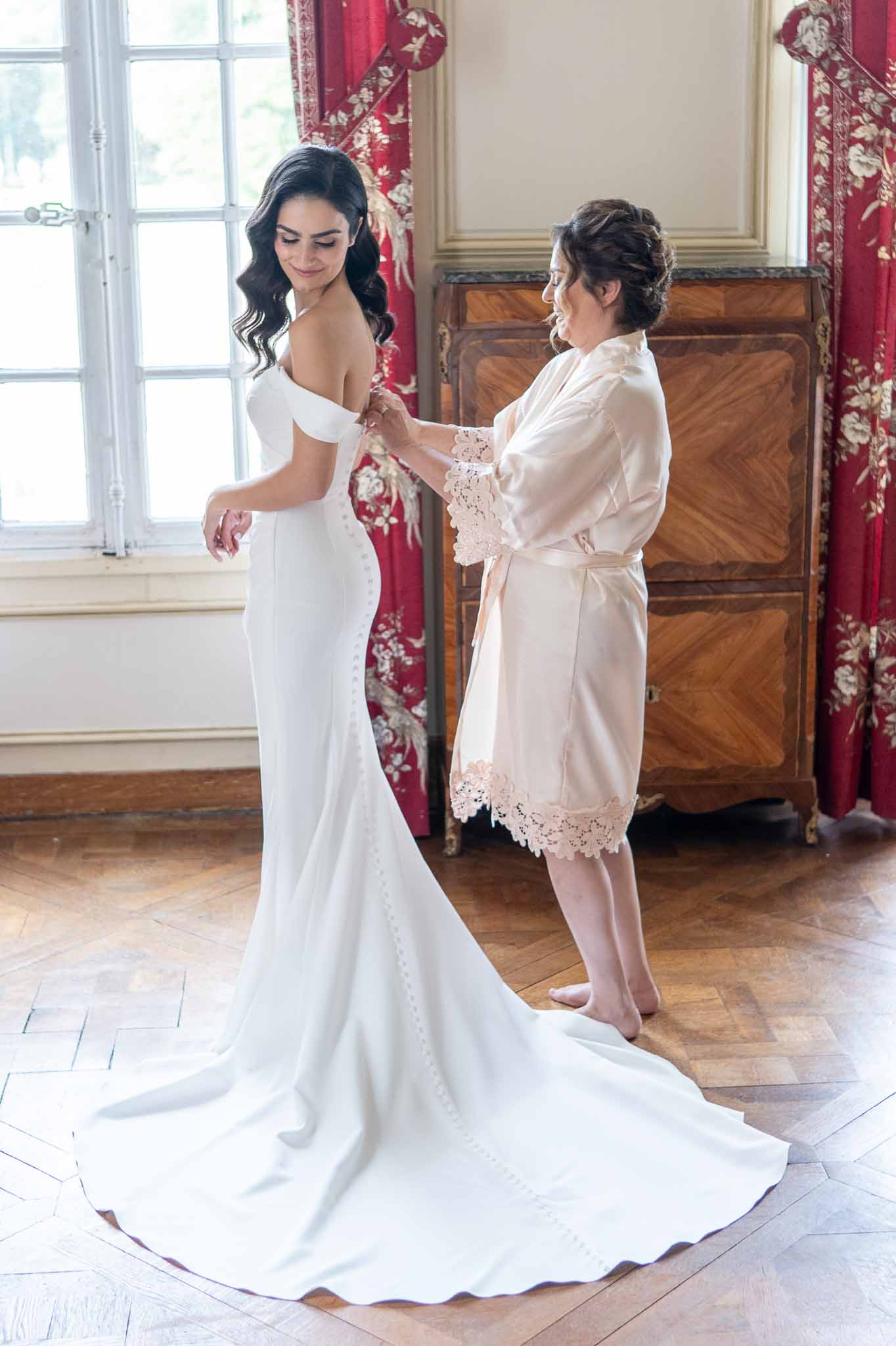 Bride being assisted with wedding dress during getting ready in classical interior room