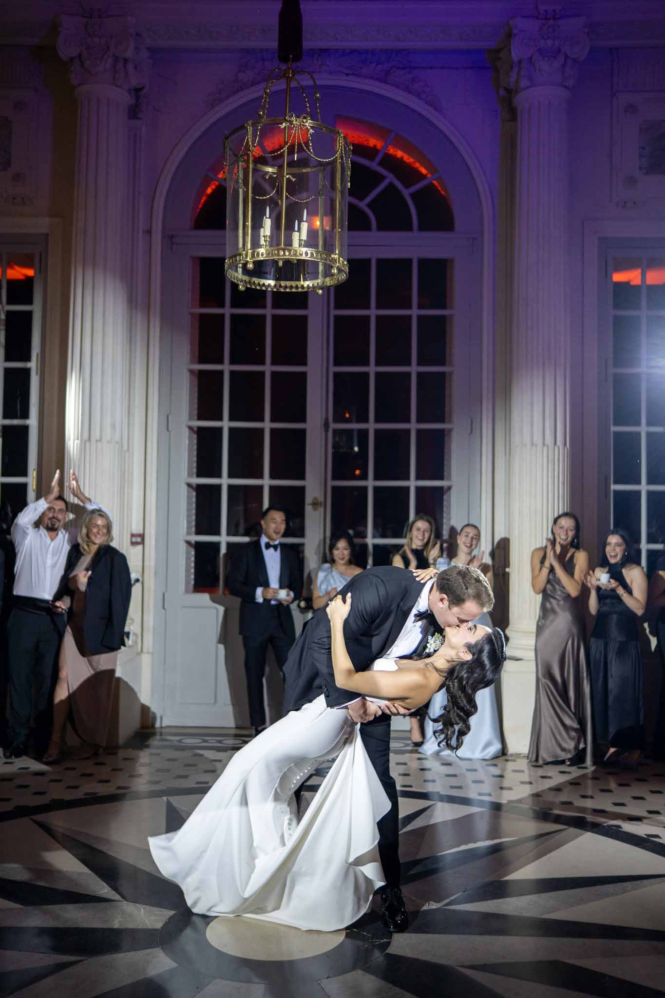 Groom dips bride during first dance in grand neoclassical ballroom with guests watching