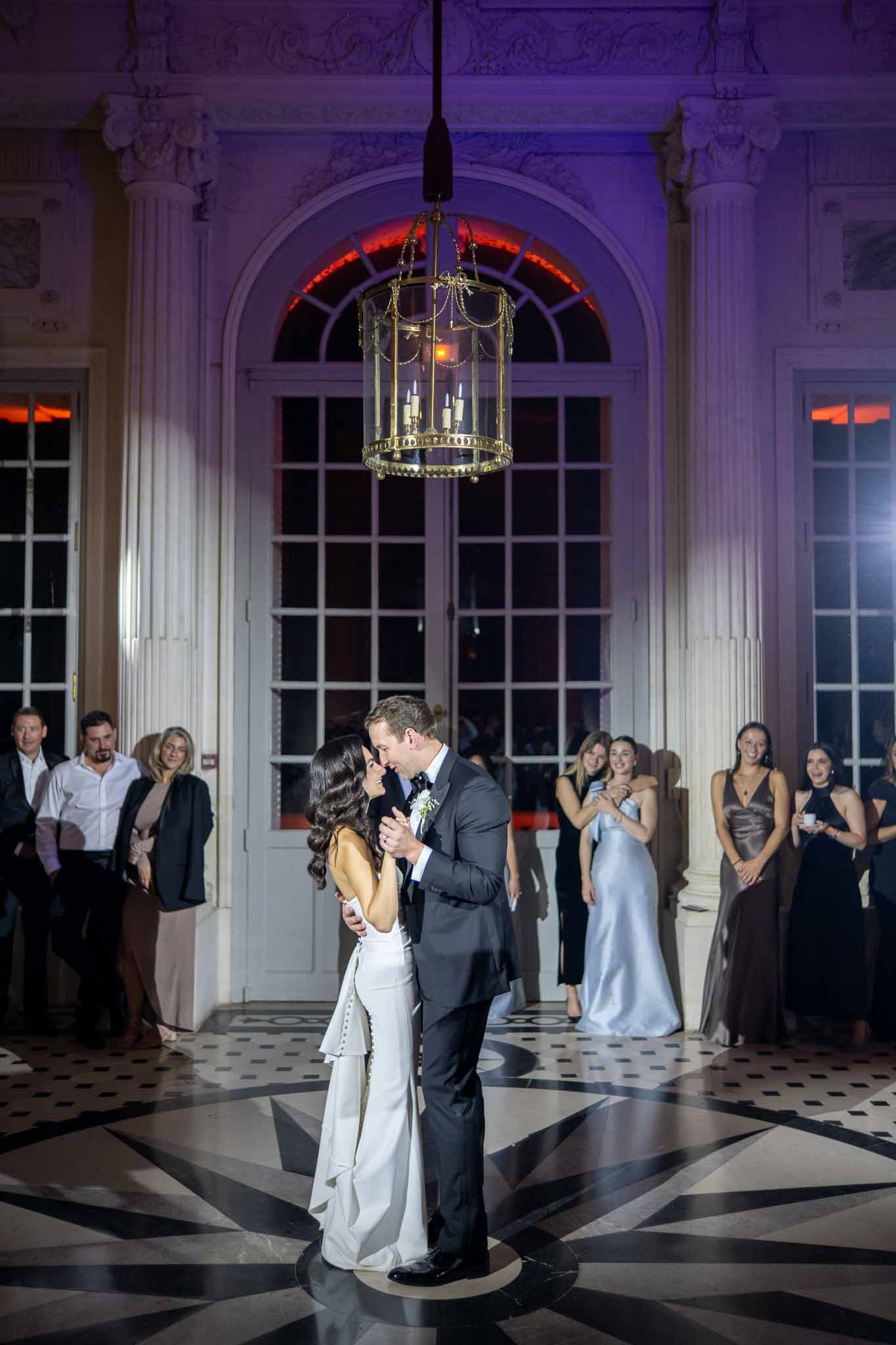 Bride and groom first dance in neoclassical ballroom with guests watching during evening reception