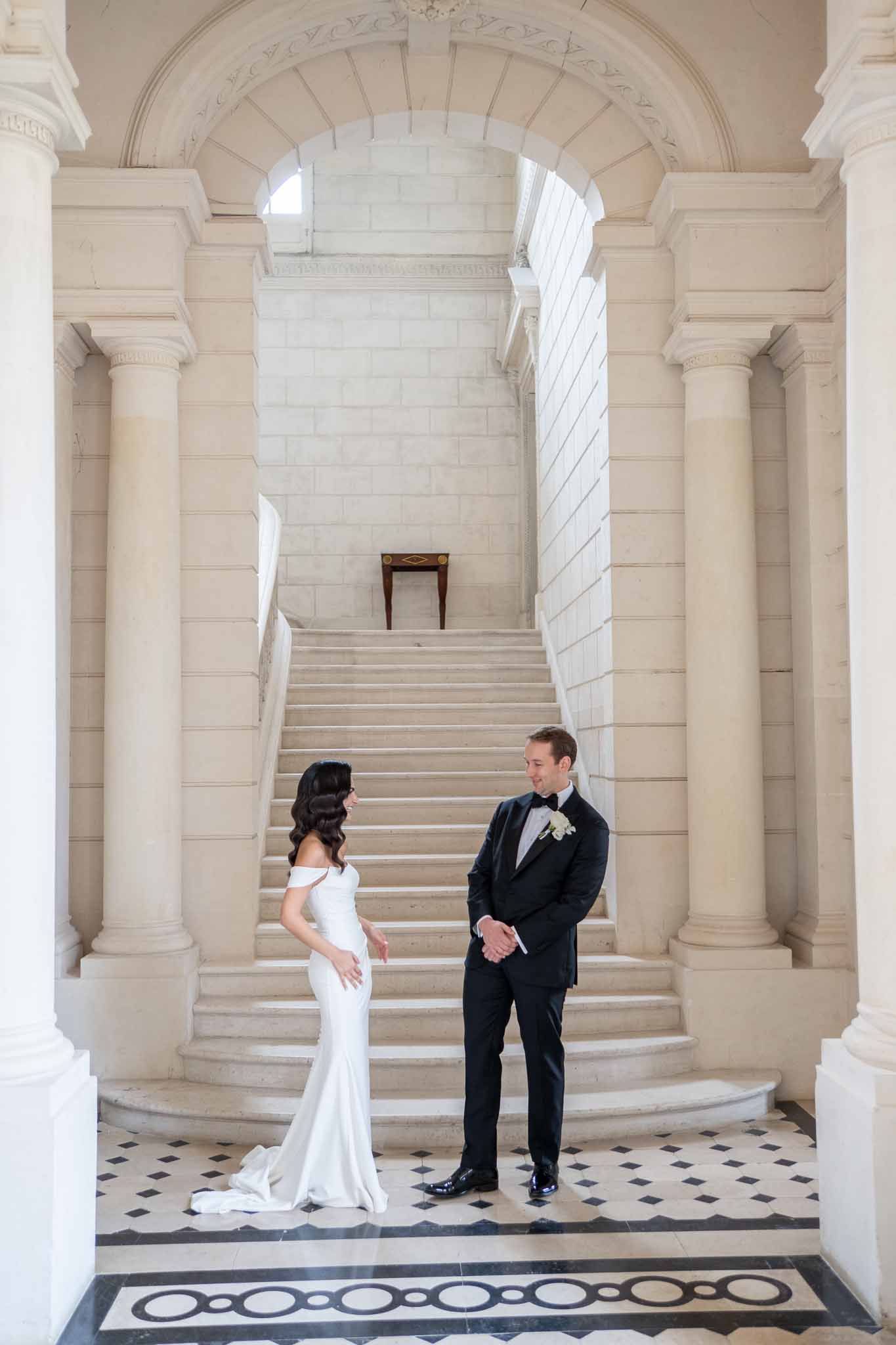 Bride and groom posing on grand staircase in classical interior with columns and arched ceiling