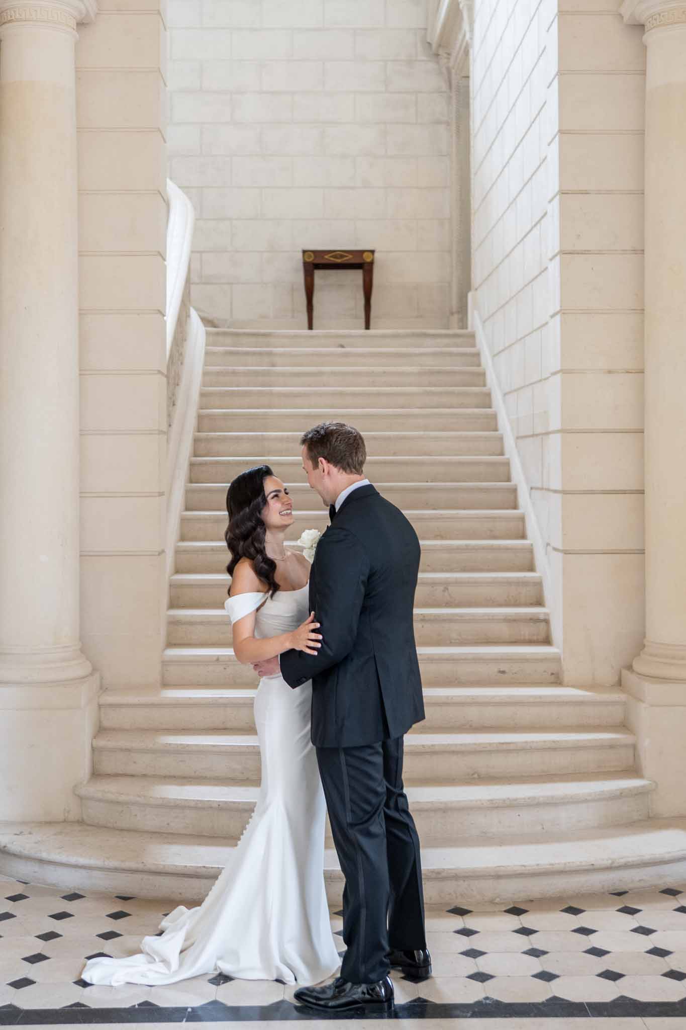 Bride and groom posing on grand staircase in classical venue with stone columns