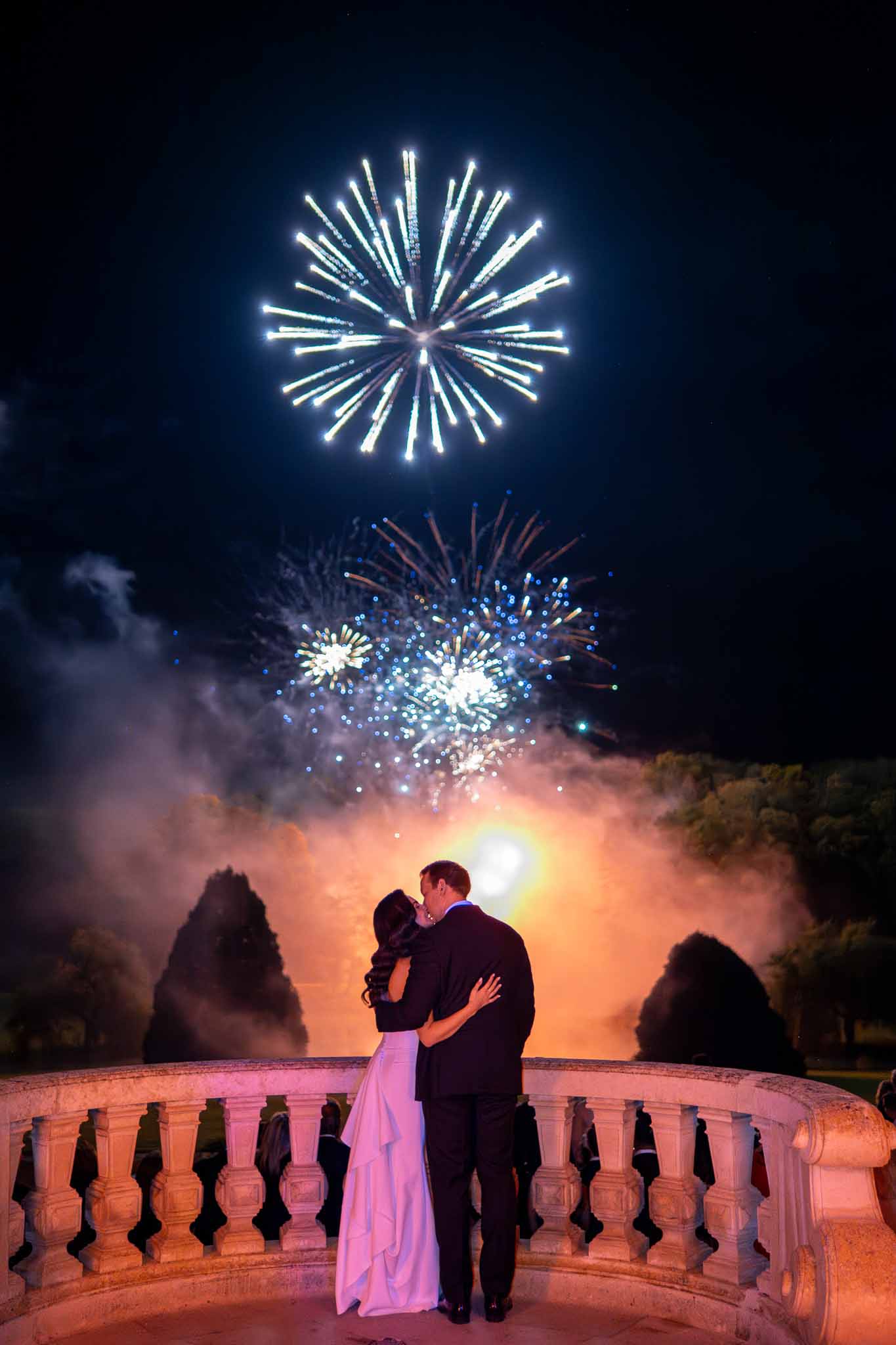 Bride and groom kissing on stone balcony during fireworks display at grand outdoor wedding venue