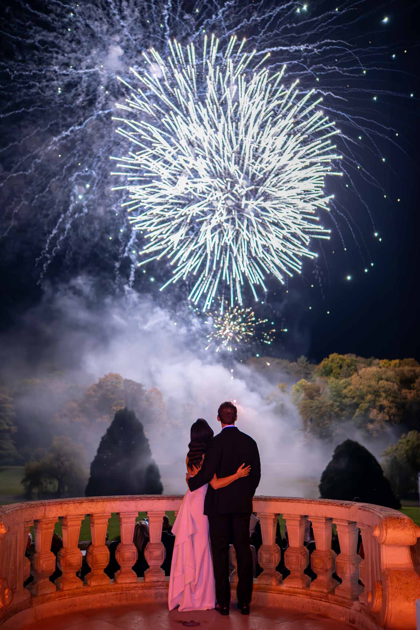 Bride and groom watching fireworks display on elegant terrace overlooking manicured gardens at night
