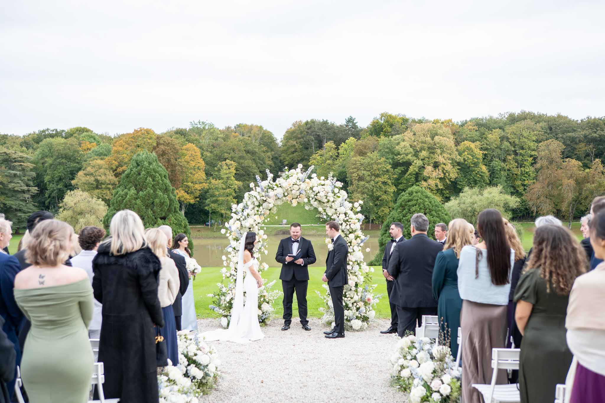 Outdoor wedding ceremony in garden setting with white floral arch and seated guests