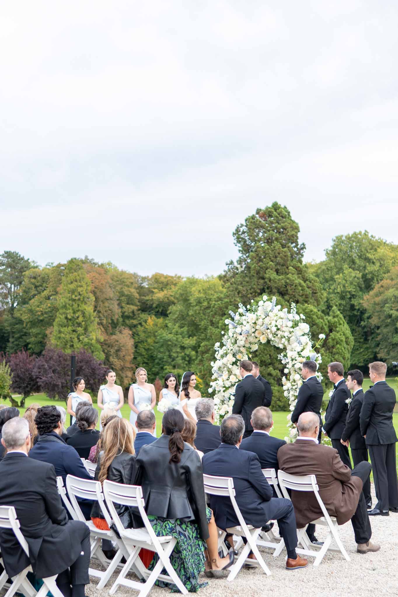 Outdoor garden wedding ceremony with white floral arch and bridal party at parkland venue