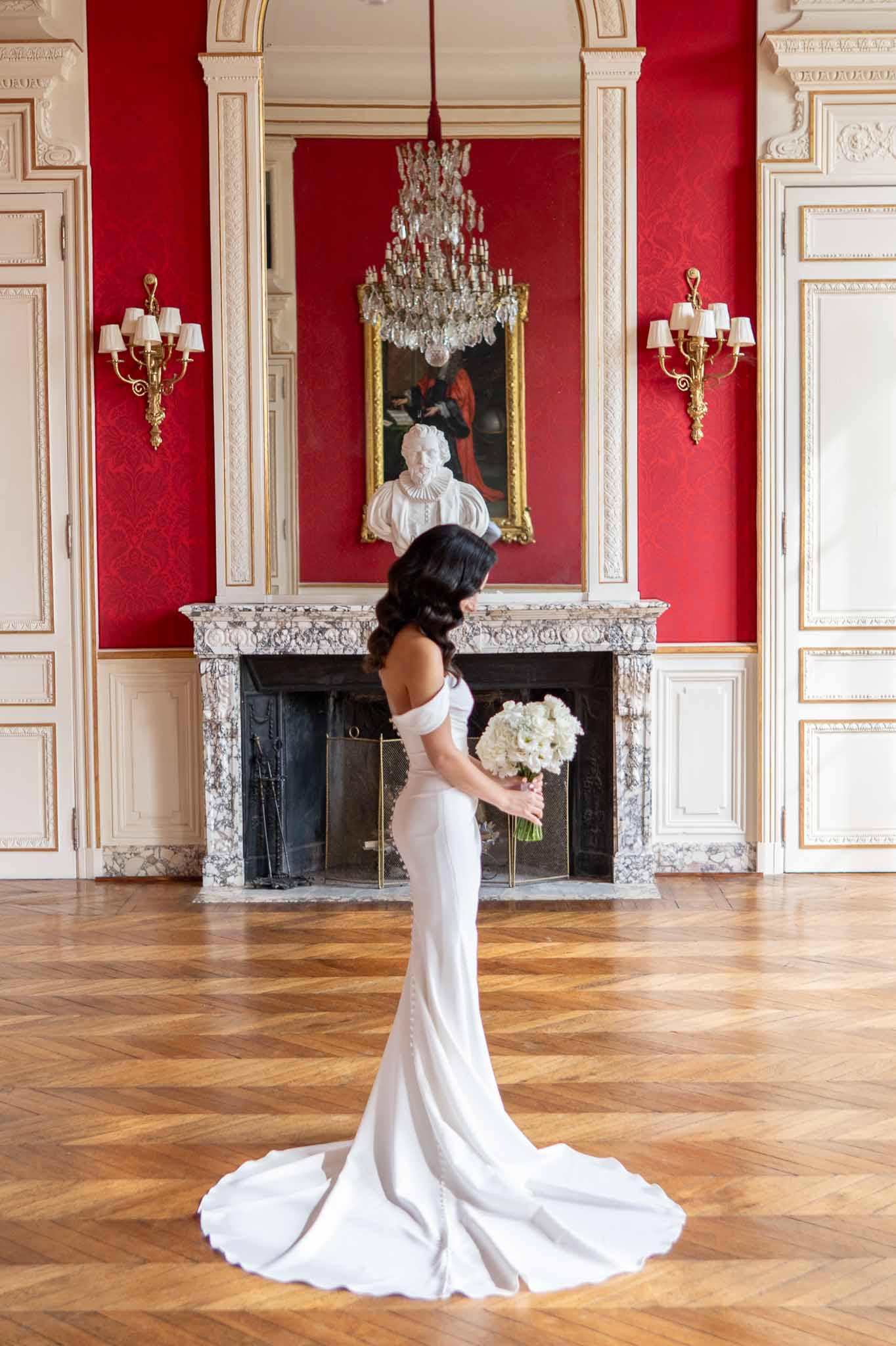 Bride in white gown with bouquet in grand neoclassical ballroom with crimson walls and crystal chandelier