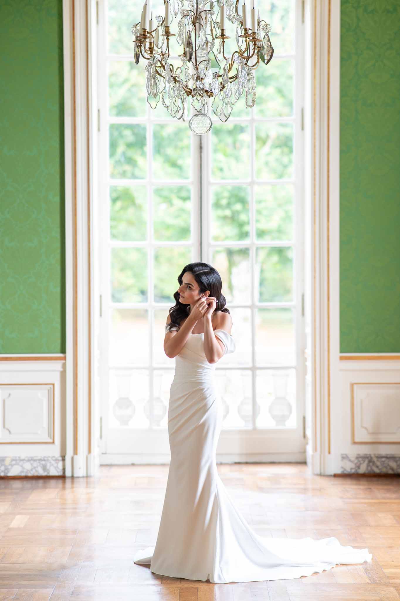 Bride in ivory silk gown adjusting earring in ornate ballroom with crystal chandelier