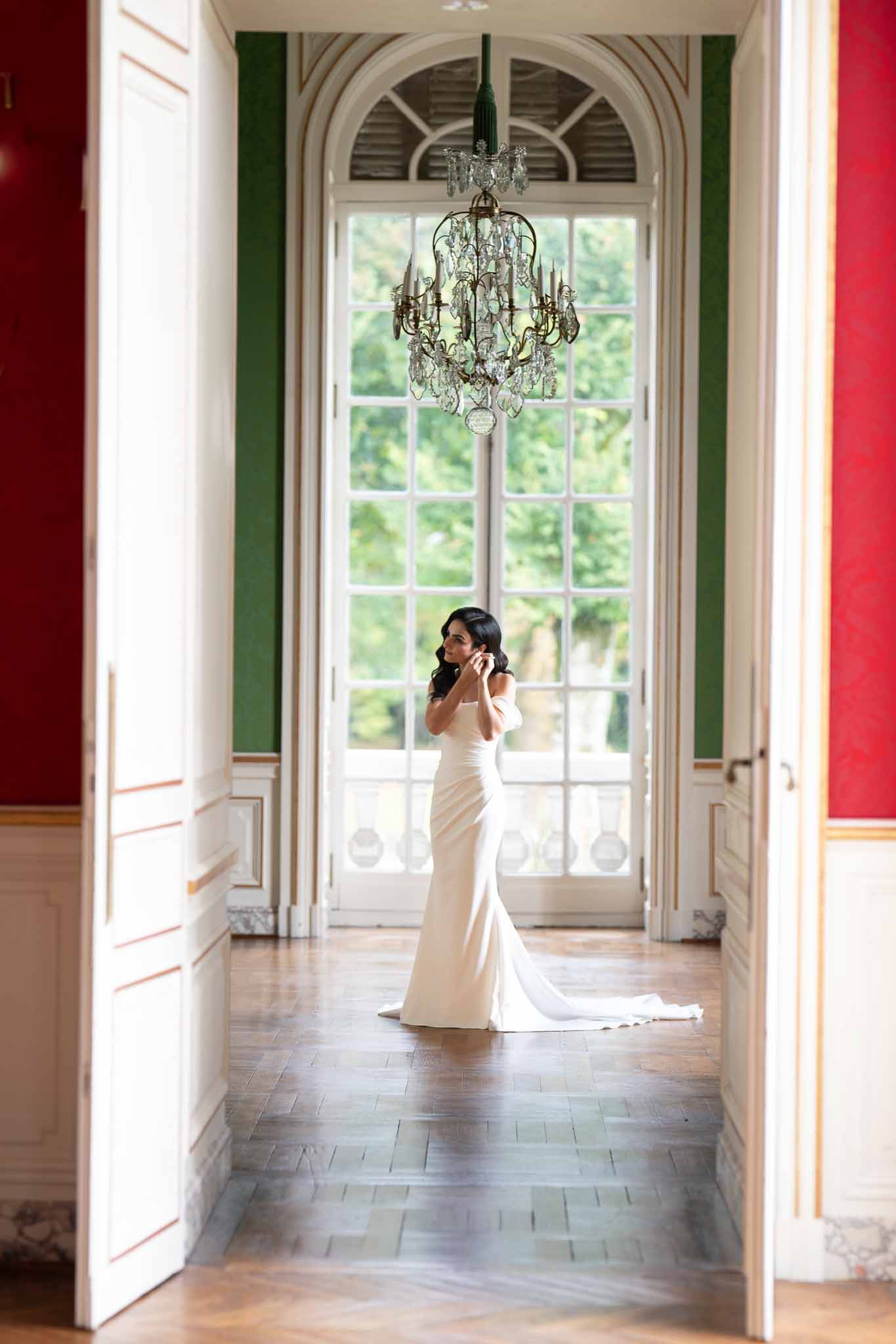 Bride in ivory crepe gown standing in classical gallery hallway with arched windows and chandelier