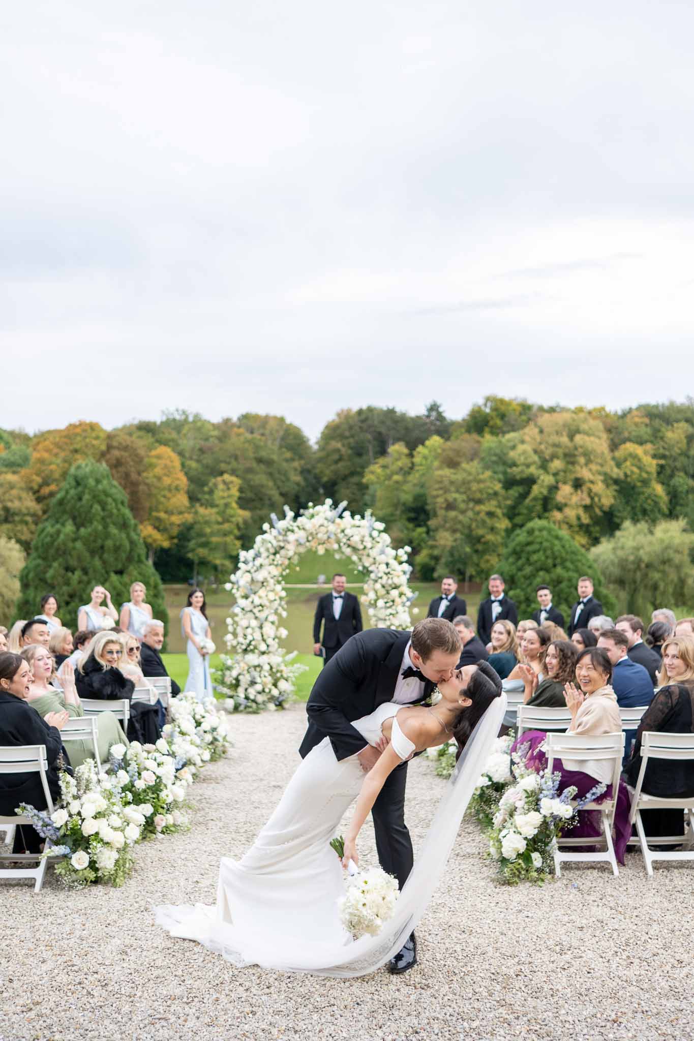 Groom dipping and kissing bride during recessional at outdoor garden ceremony with white floral arch
