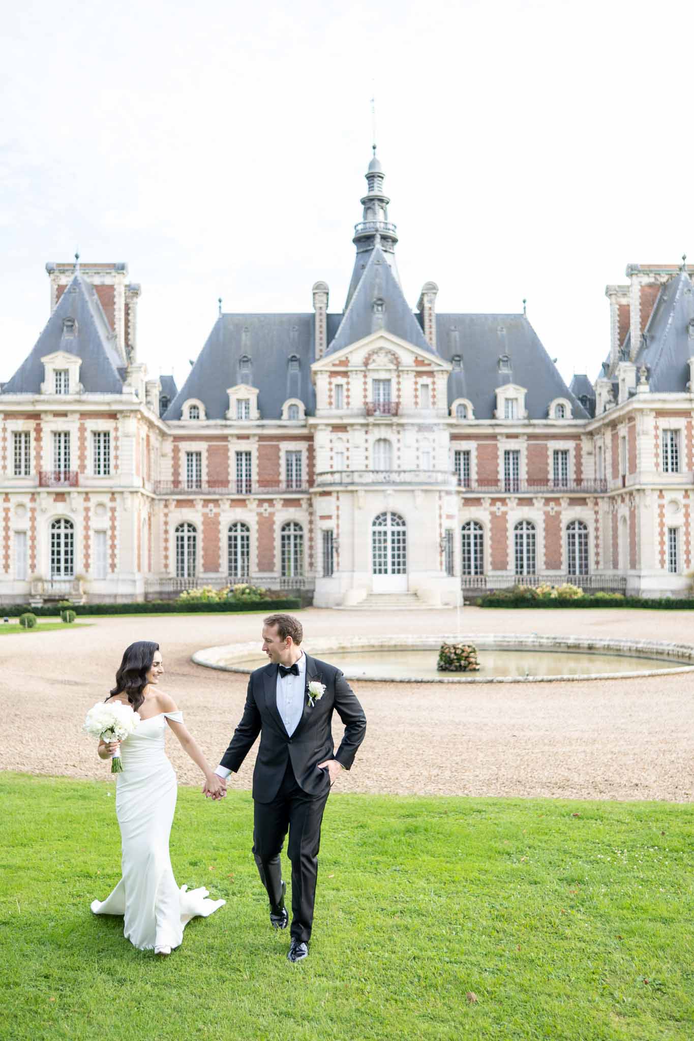 Bride and groom walking hand-in-hand across courtyard lawn in front of grand French château