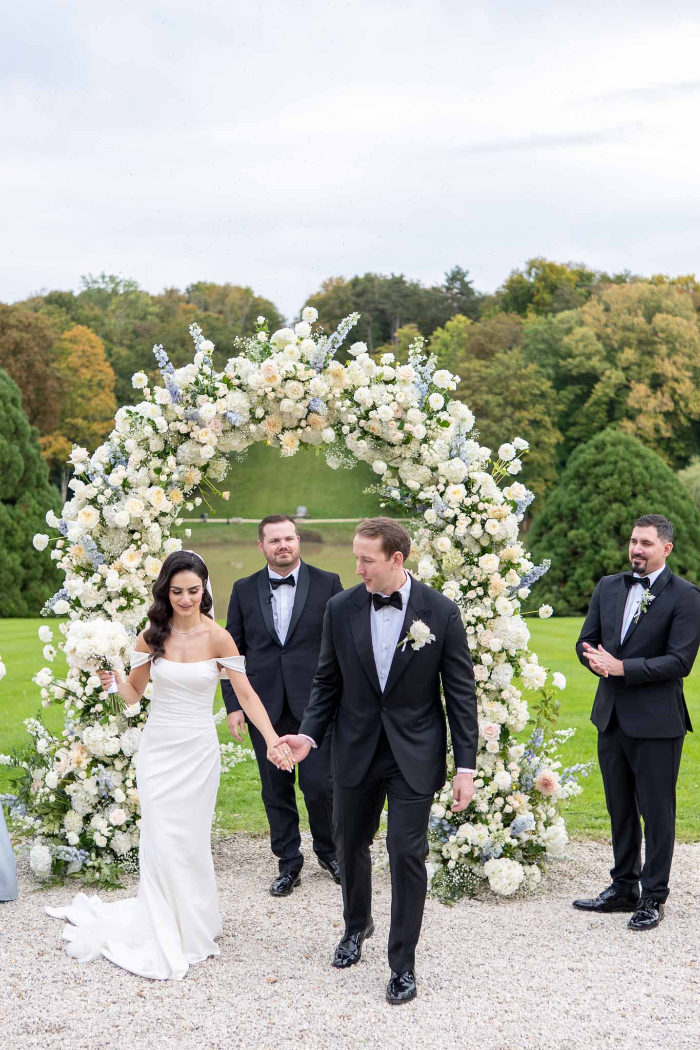 Bride and groom walking beneath floral arch during outdoor wedding ceremony recession