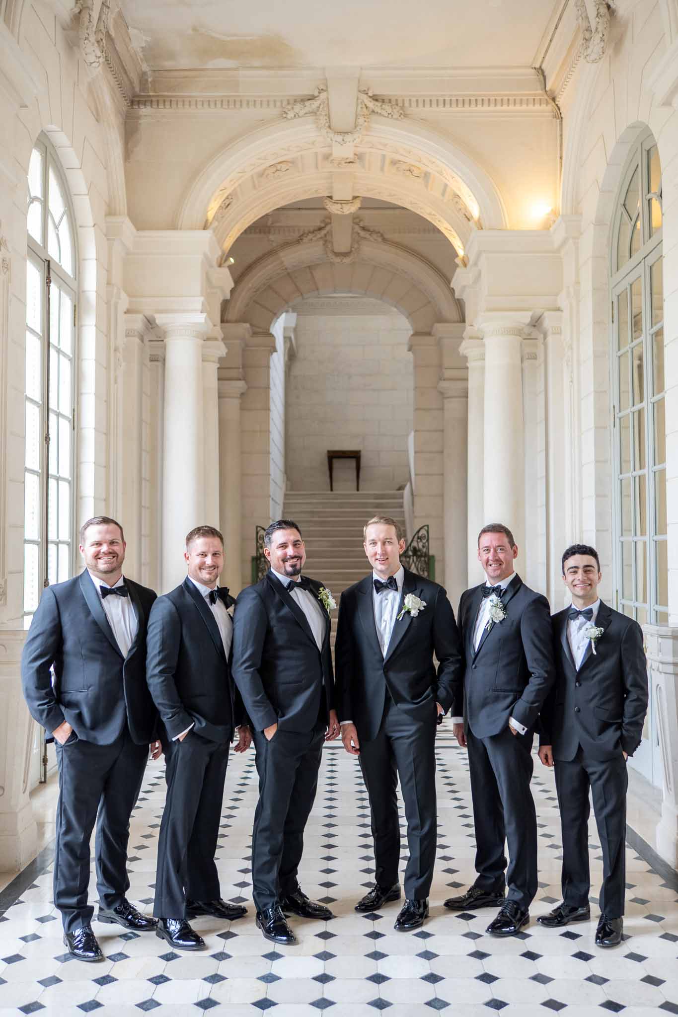 Groom and groomsmen in navy tuxedos standing in classical stone corridor with vaulted archways