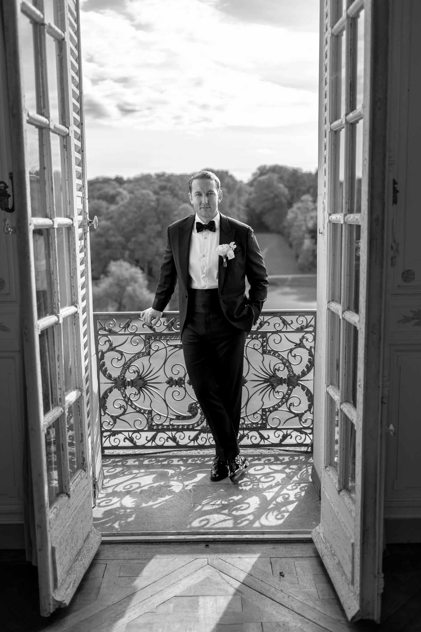 Groom in black tuxedo standing on wrought-iron balcony at French chateau wedding venue