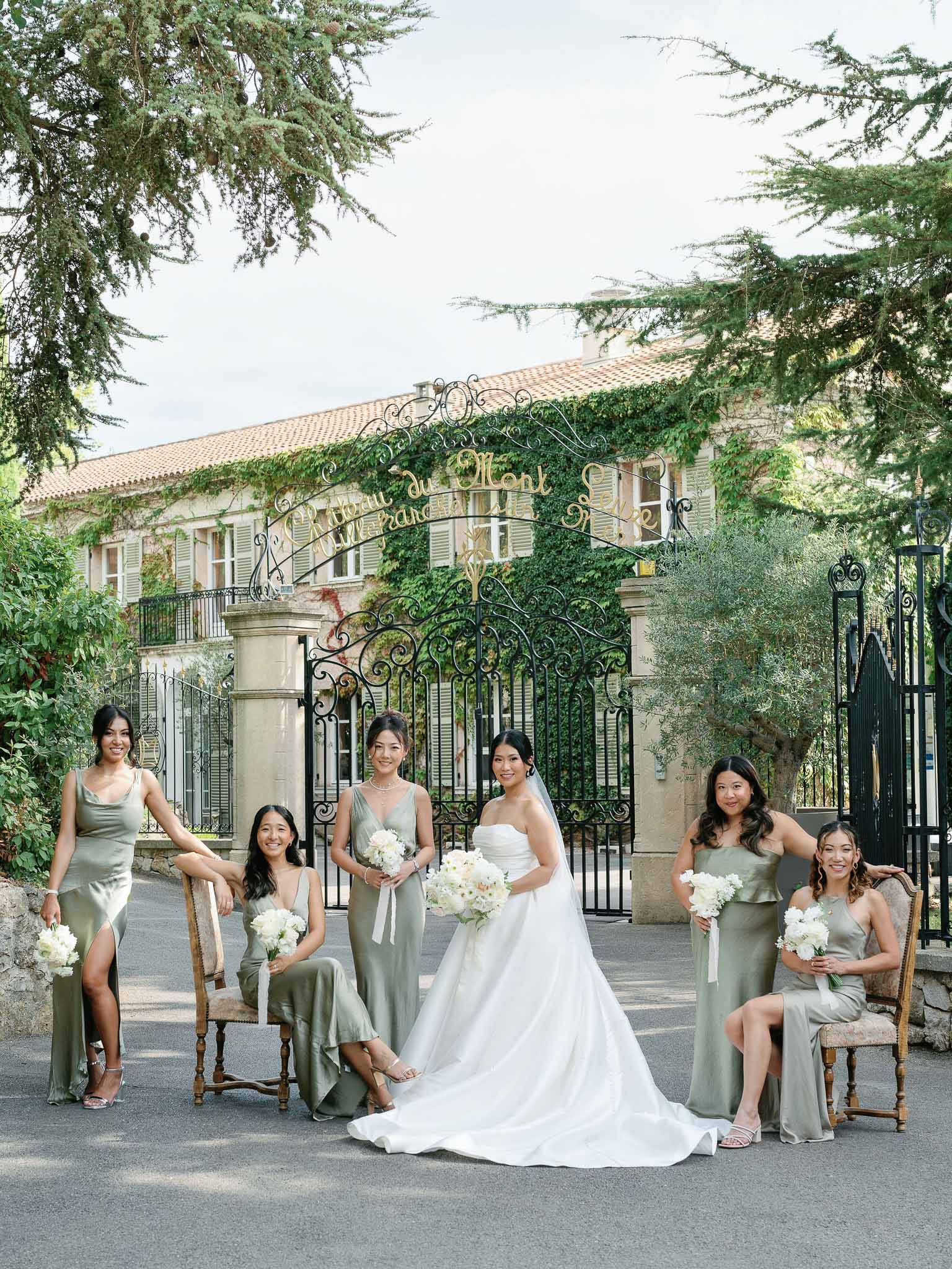 Bride and bridesmaids in sage green dresses posing with bouquets in front of European villa with iron gates