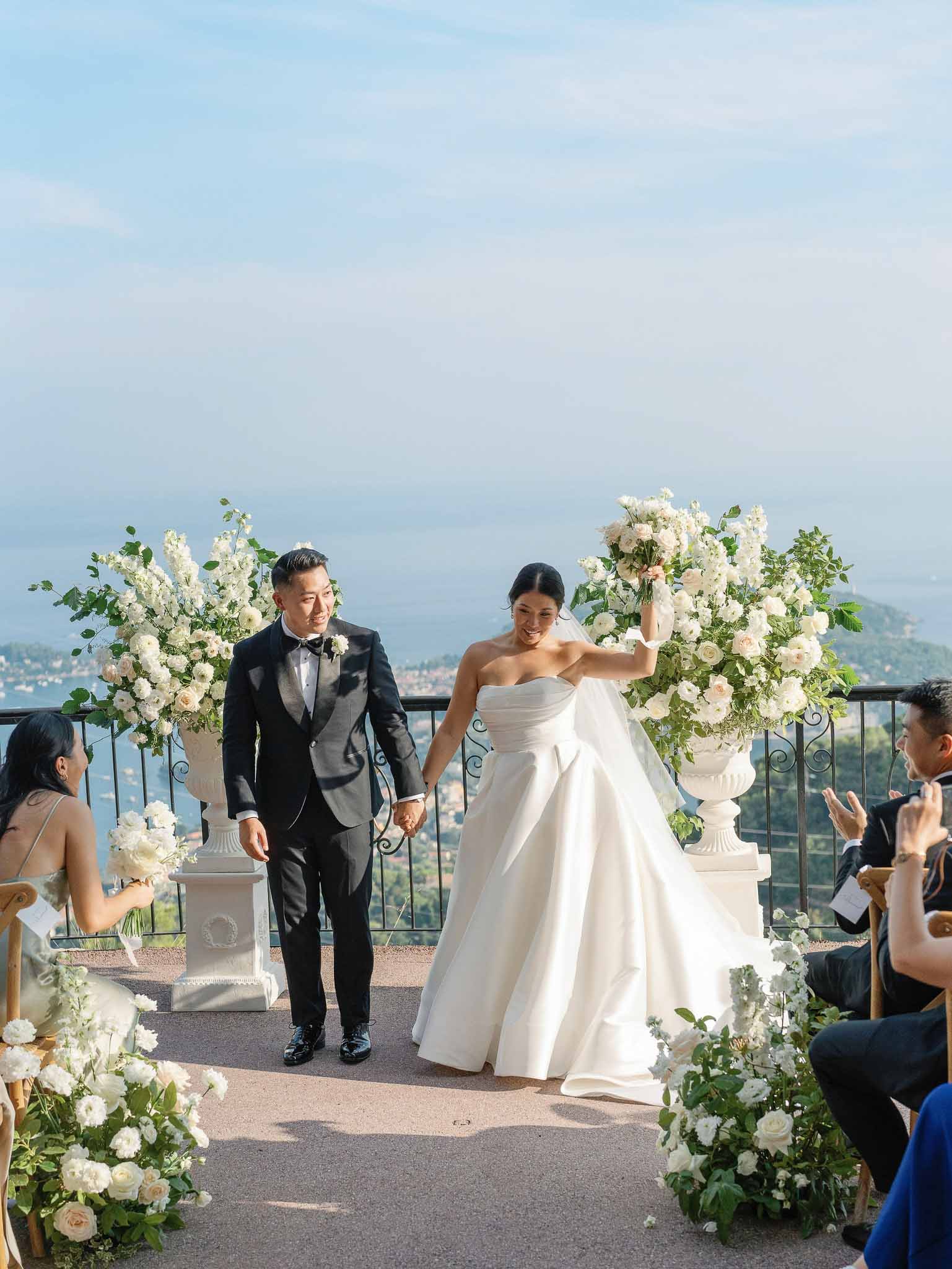 Bride and groom walking down outdoor ceremony aisle at mountain terrace venue with white floral arrangements