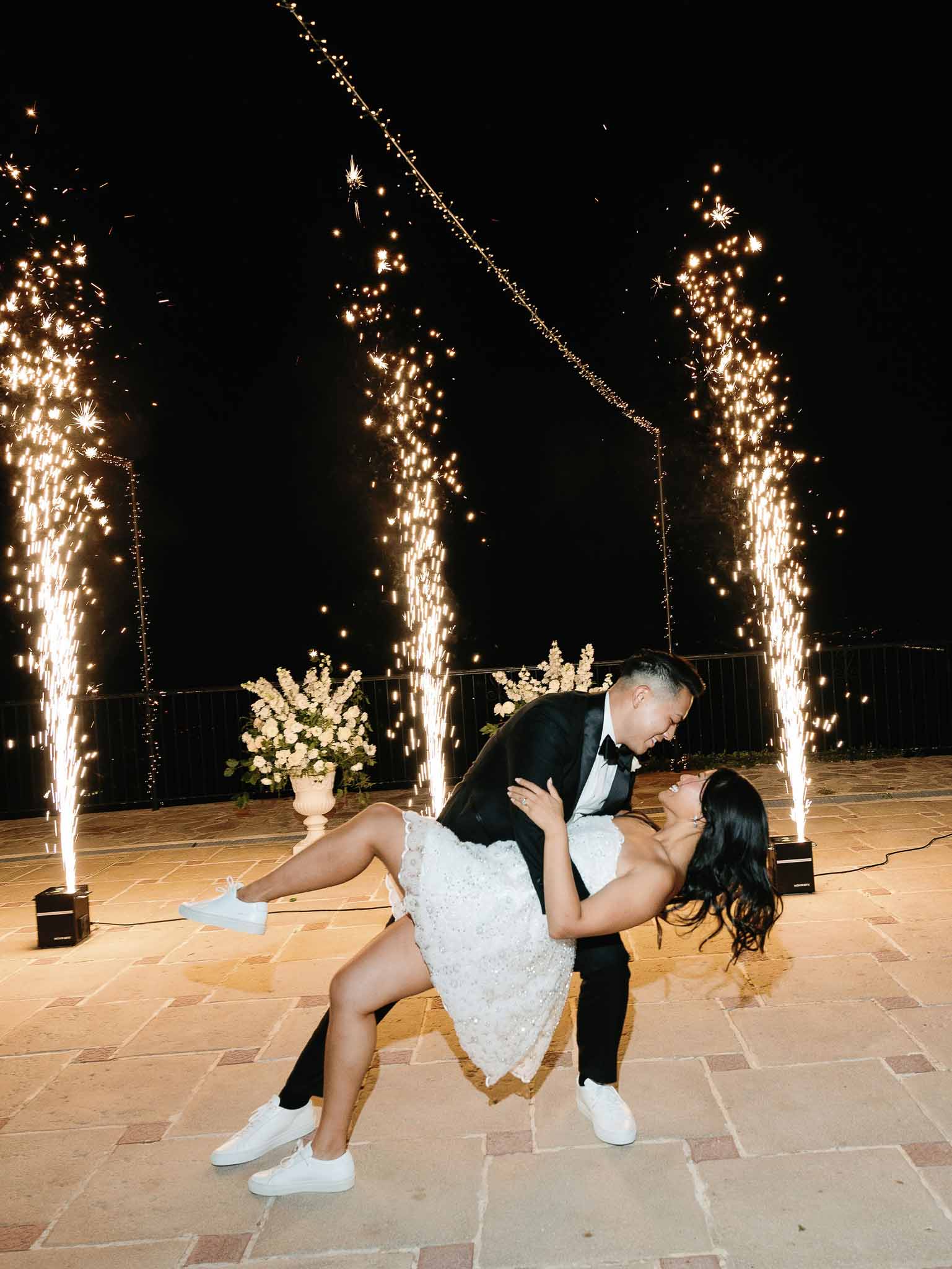 Groom lifts bride during first dance on outdoor terrace with fairy lights at night wedding reception