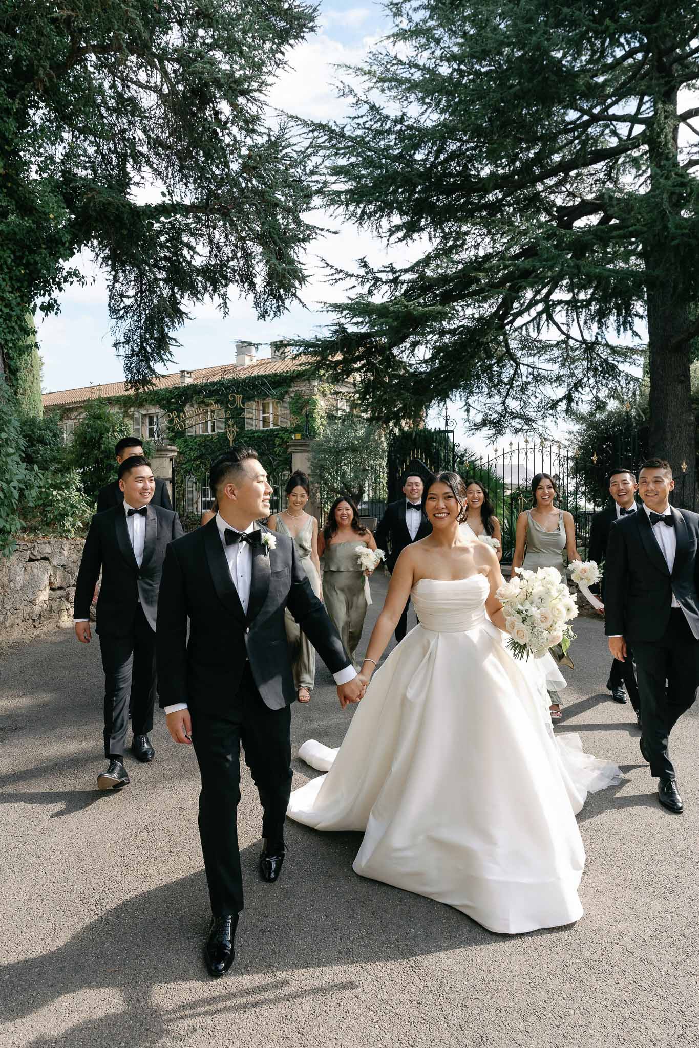 Bride and groom walking down tree-lined driveway with bridal party at European estate wedding