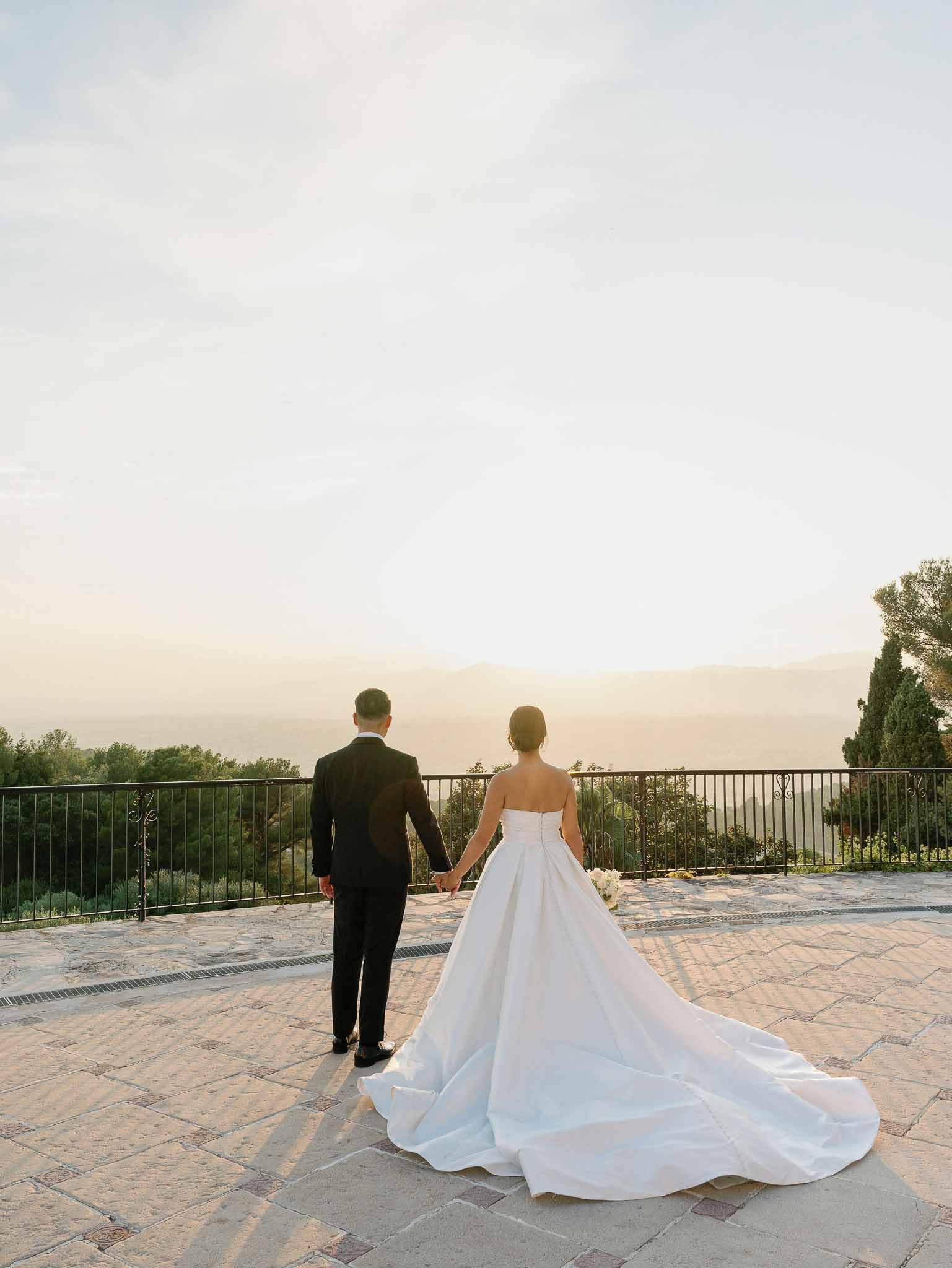 Bride and groom holding hands on stone terrace overlooking landscape at golden hour