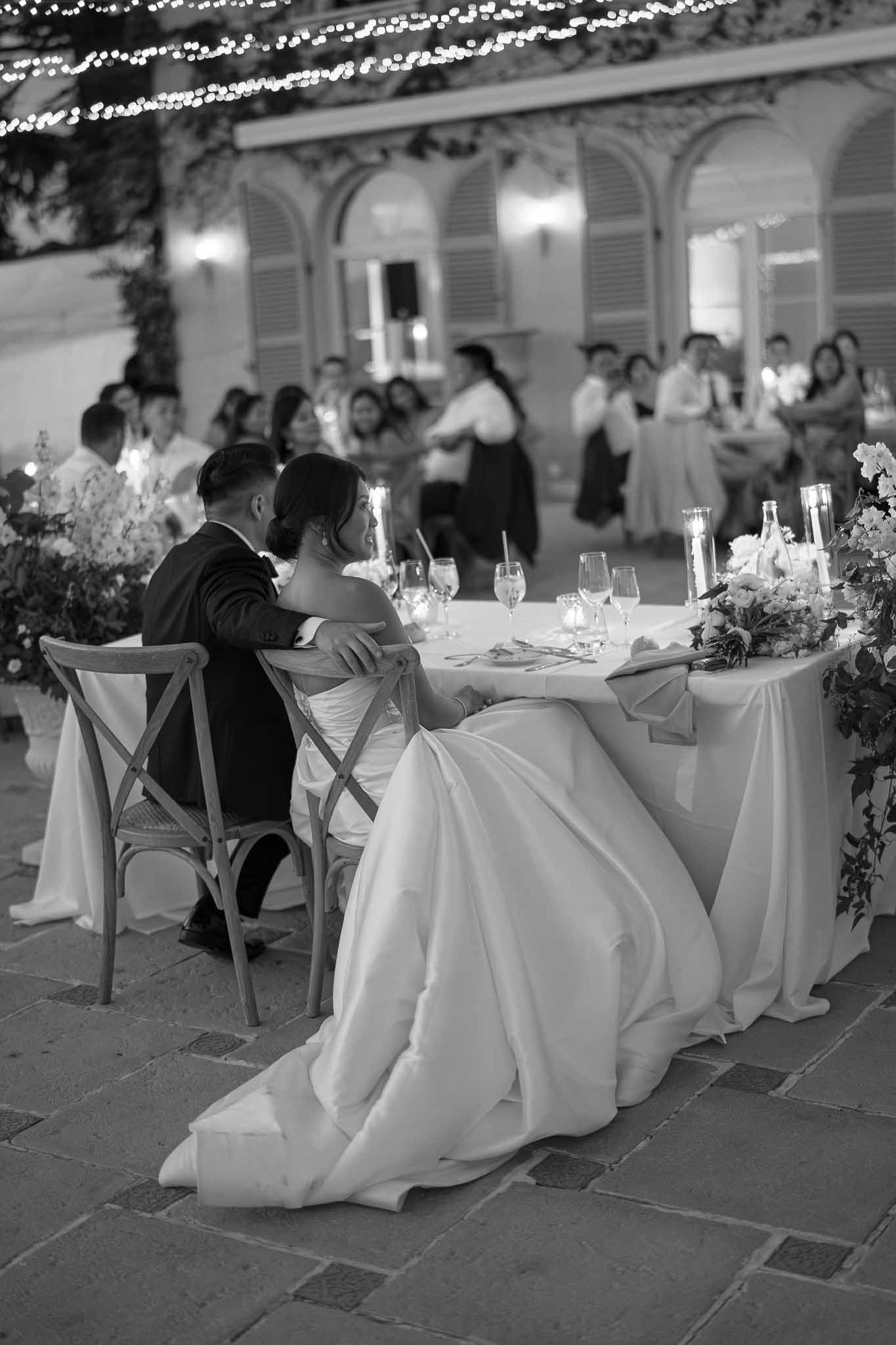 Bride and groom at sweetheart table during outdoor courtyard reception with dancing guests in background