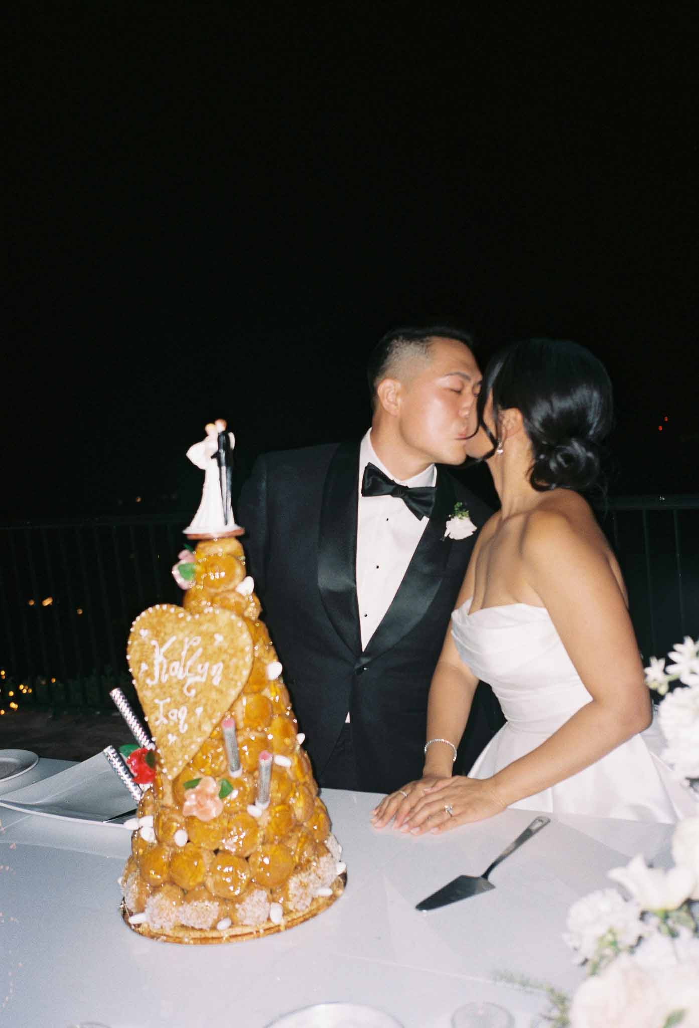 Bride and groom kissing during cake cutting with croquembouche at indoor wedding reception