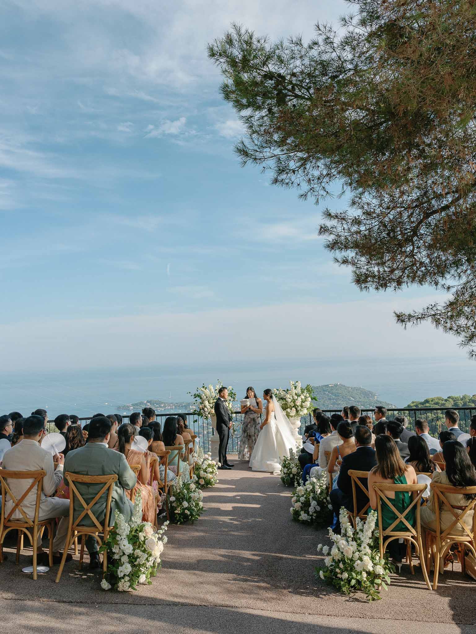 Outdoor wedding ceremony on clifftop terrace overlooking Mediterranean Sea with guests in wooden chairs