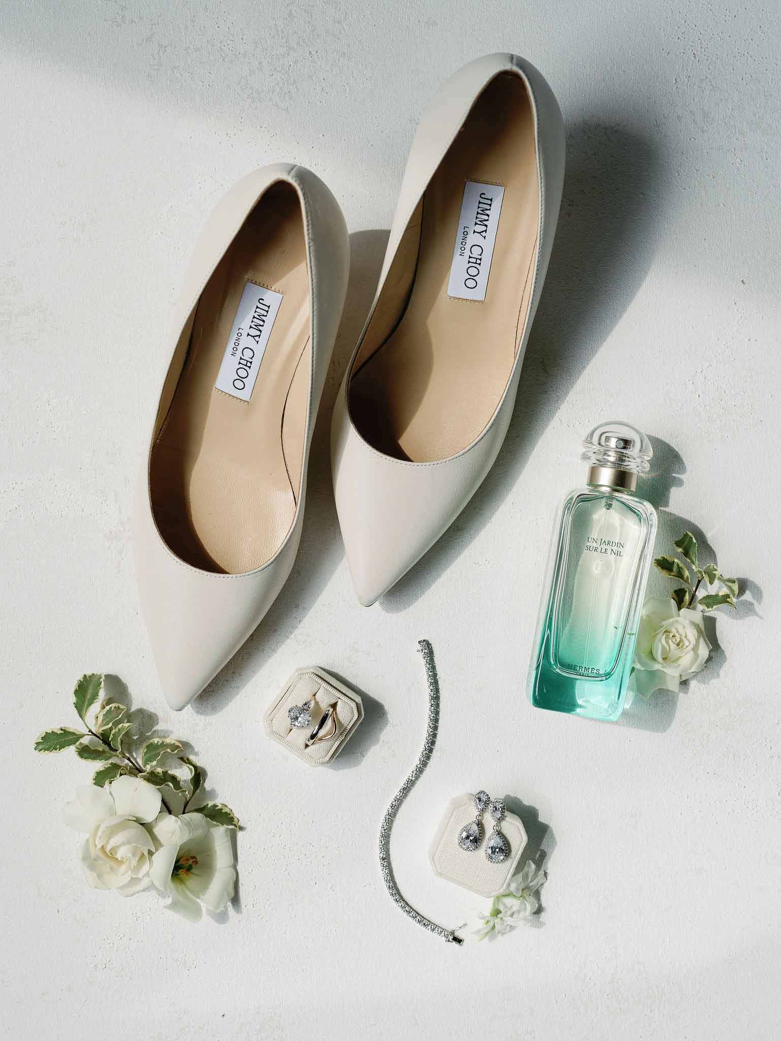 Bridal accessories flat lay with Jimmy Choo shoes, rings, and white roses on textured surface