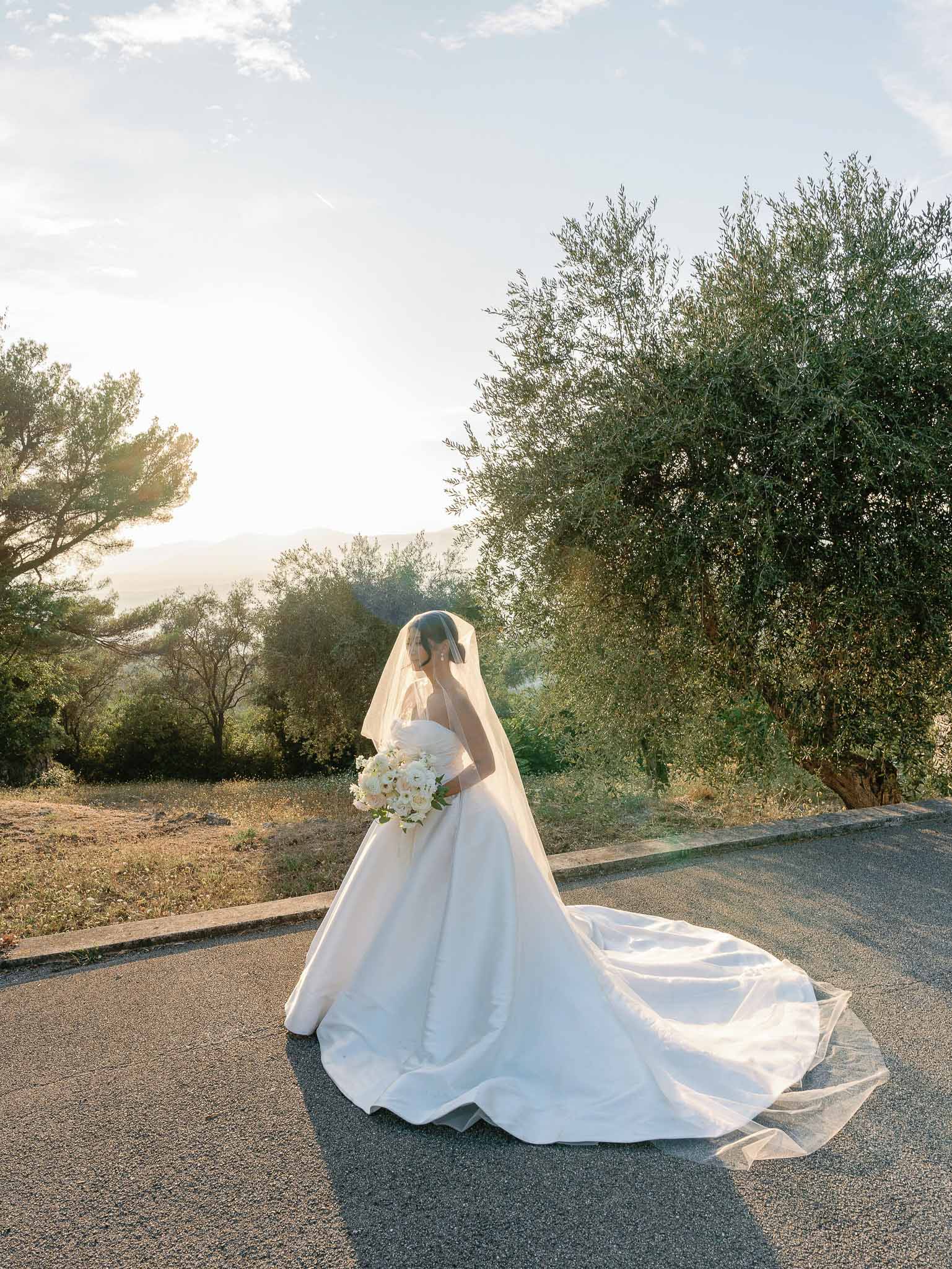 Bride in ivory ball gown standing on olive tree-lined road in Mediterranean countryside