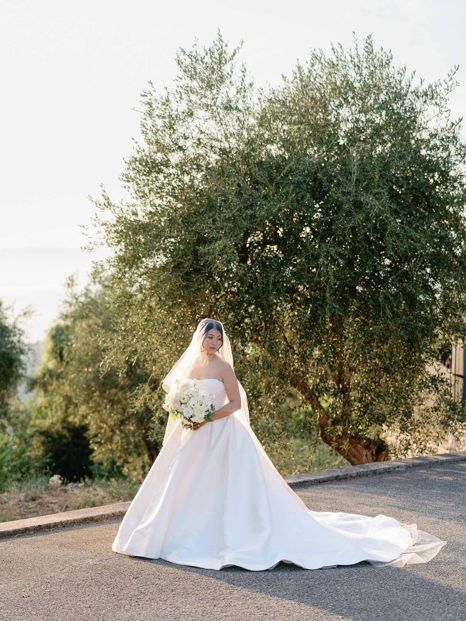 Bride in ivory ball gown with bouquet standing on tree-lined rural road for portrait