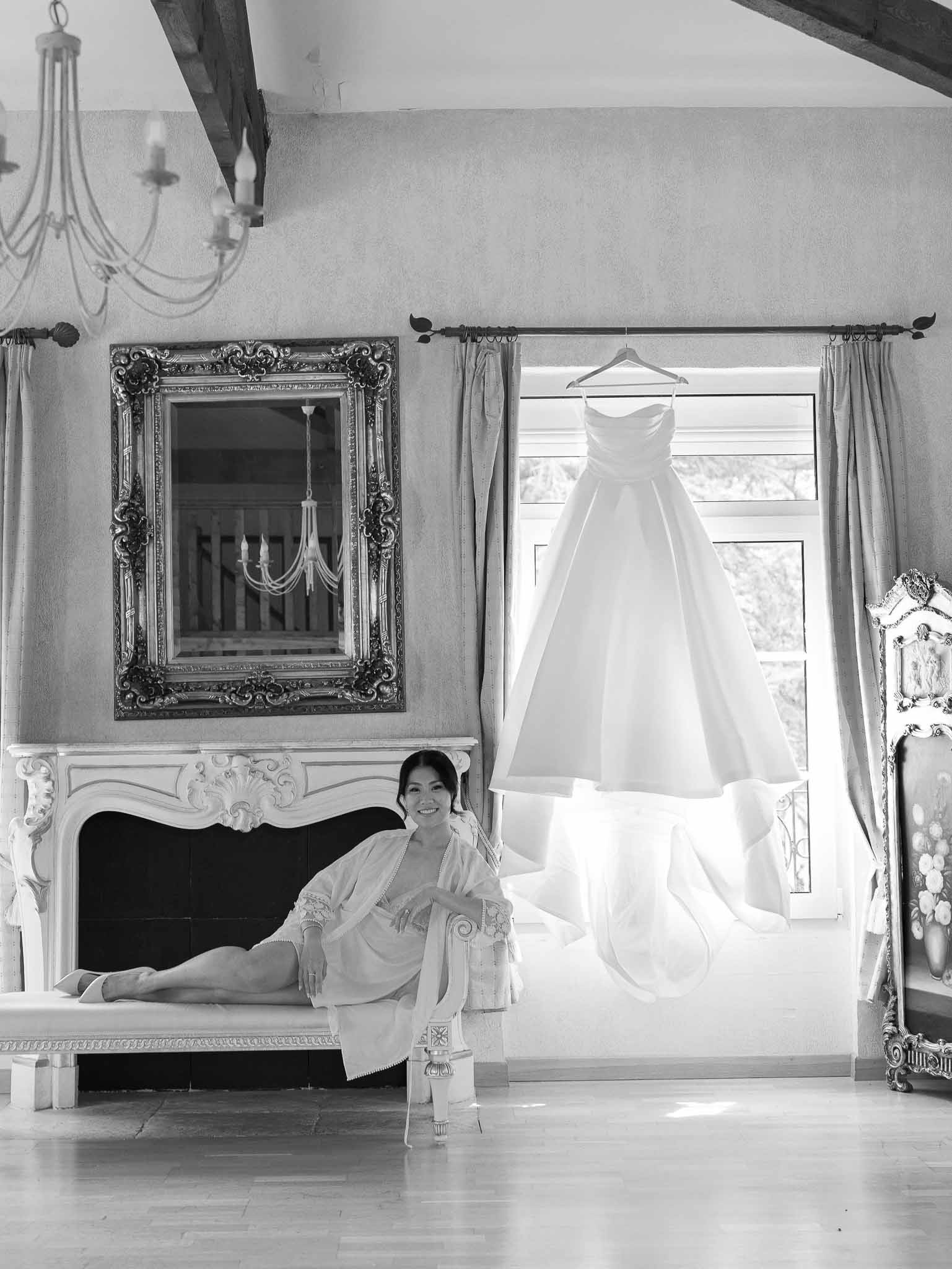 Bride in robe sitting on fireplace mantel with wedding dress hanging by window in ornate indoor space