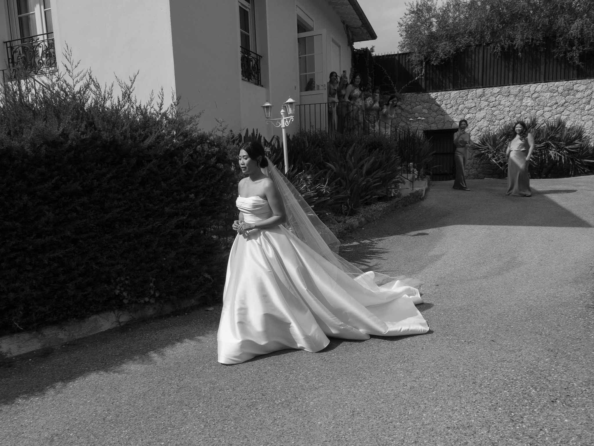 Bride in strapless ball gown and veil posing in courtyard of white residence with guests on terrace