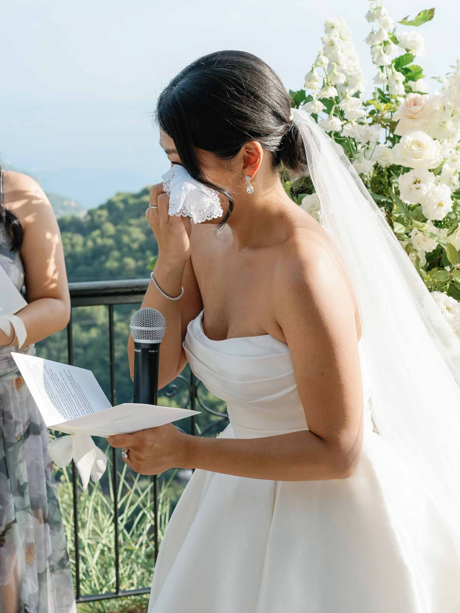 Bride giving emotional speech during outdoor ceremony with floral backdrop and landscape views