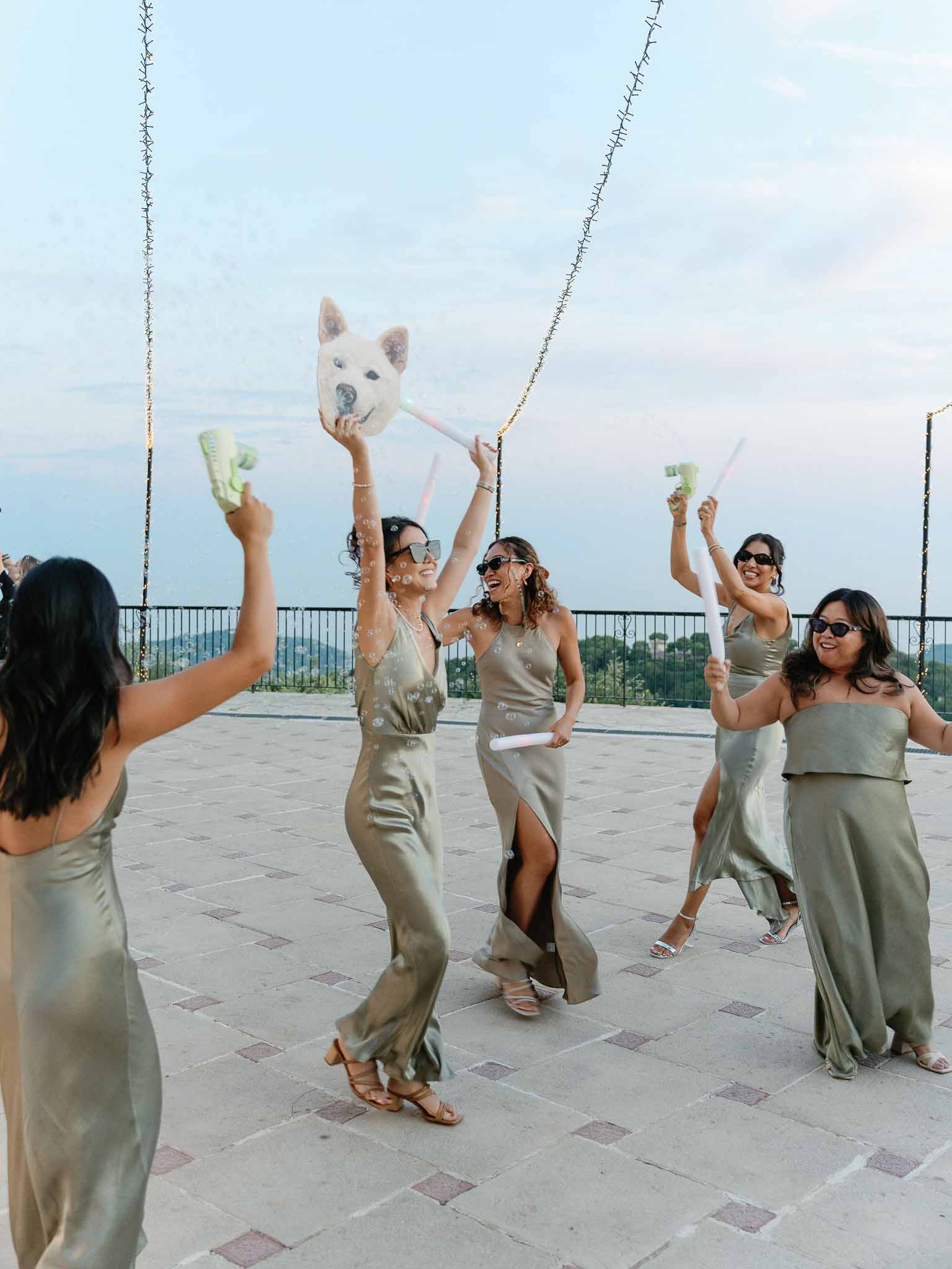 Bridesmaids in sage green dresses celebrating with confetti poppers on terrace overlooking countryside